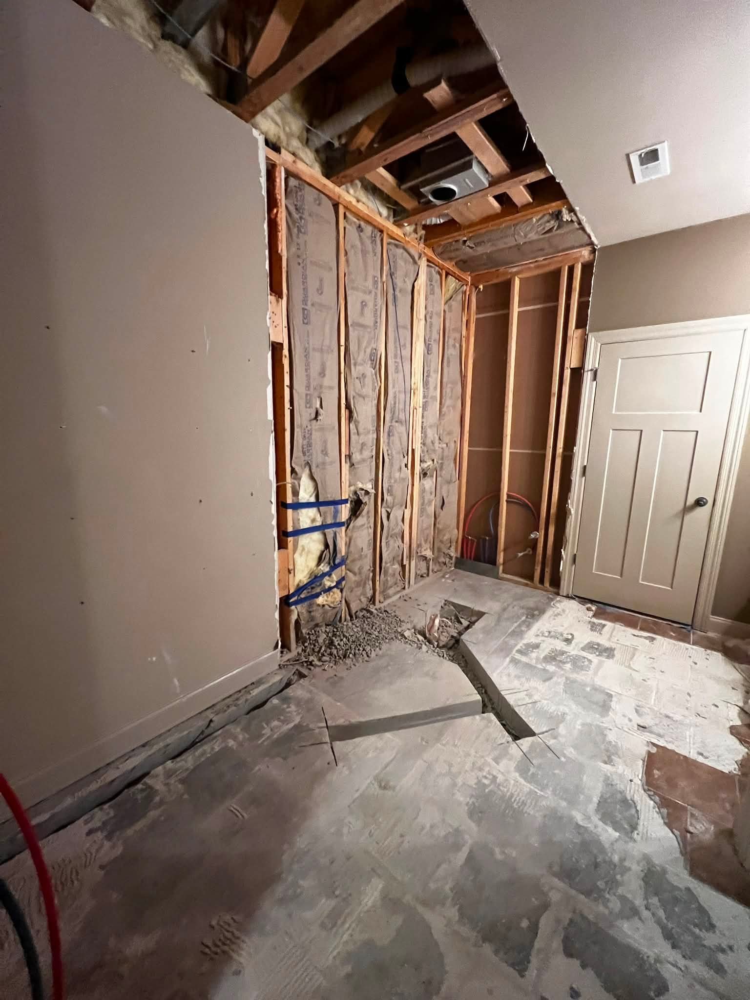 A room undergoing renovation with exposed wall framing, insulation, and drywall missing in sections. The floor is partially stripped, revealing concrete and construction debris.