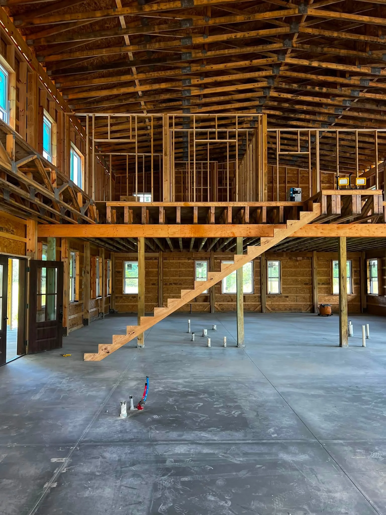 Interior of a house under construction with unfinished wooden framing, exposed beams, and partially installed plumbing. The space has large windows, a sliding door, and a concrete floor.