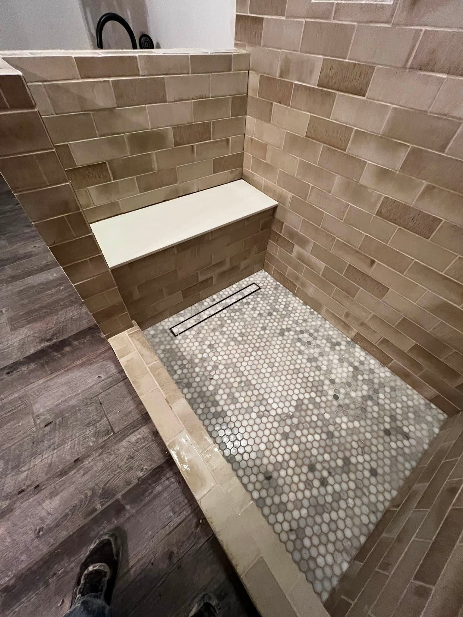 A walk-in shower area with beige brick tiles on the wall and small white hexagonal tiles on the floor. A white bench is built into the wall, and there is a black drain cover on the floor. Part of a person's feet wearing sneakers are visible at the bo