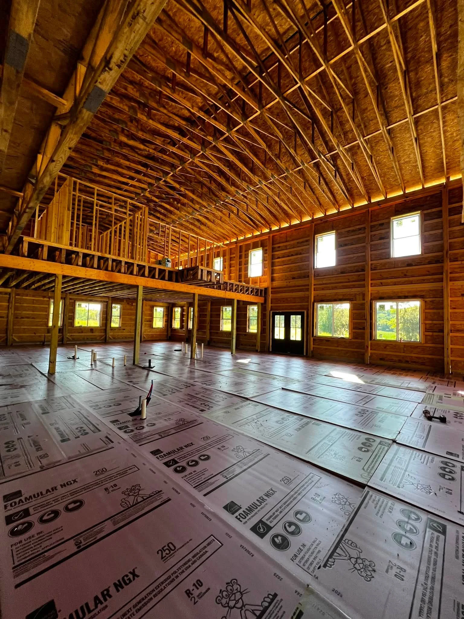 Interior of a building under construction, with a wooden framework for the roof, multiple window openings, and a floor covered with pink foam insulation sheets for a building foundation or basement.
