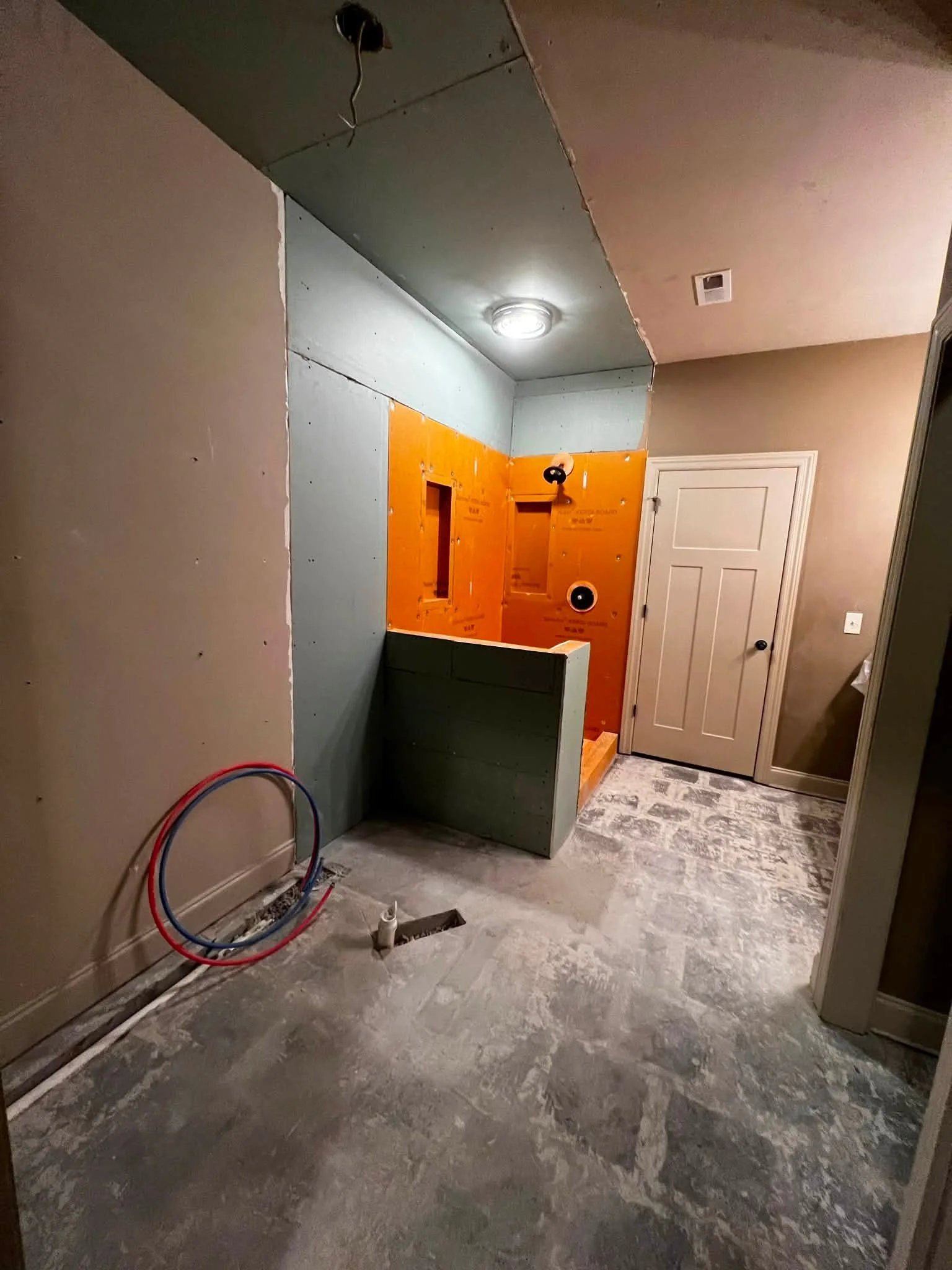 Interior room under renovation with exposed electrical wiring, drywall, and flooring. Wall with wiring and an outlet box, and an unfinished ceiling with light fixture. Closed door visible on the right.