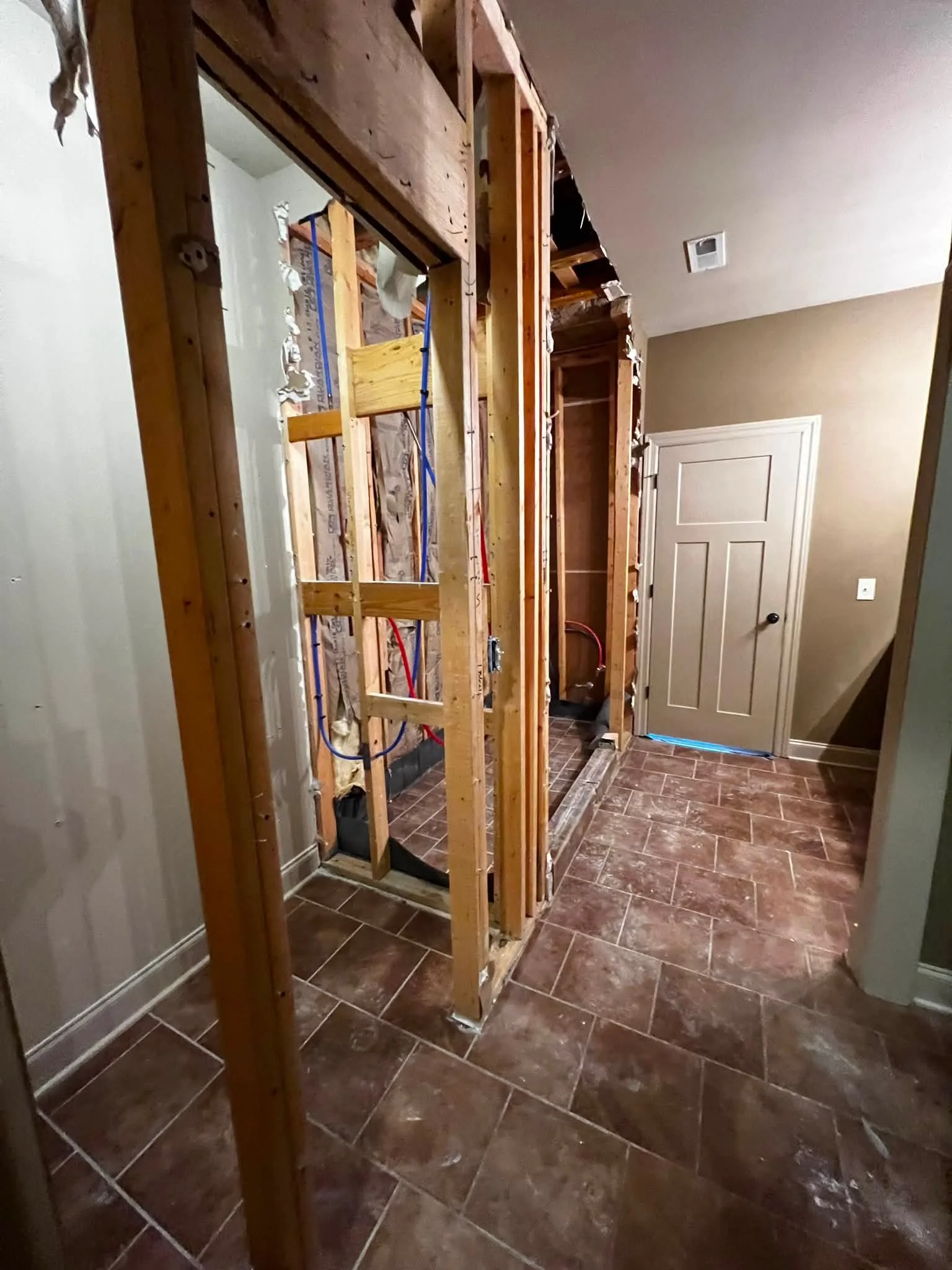 The room under renovation with exposed wooden framing, partially removed wall, electrical wiring, and brown tiled floor.