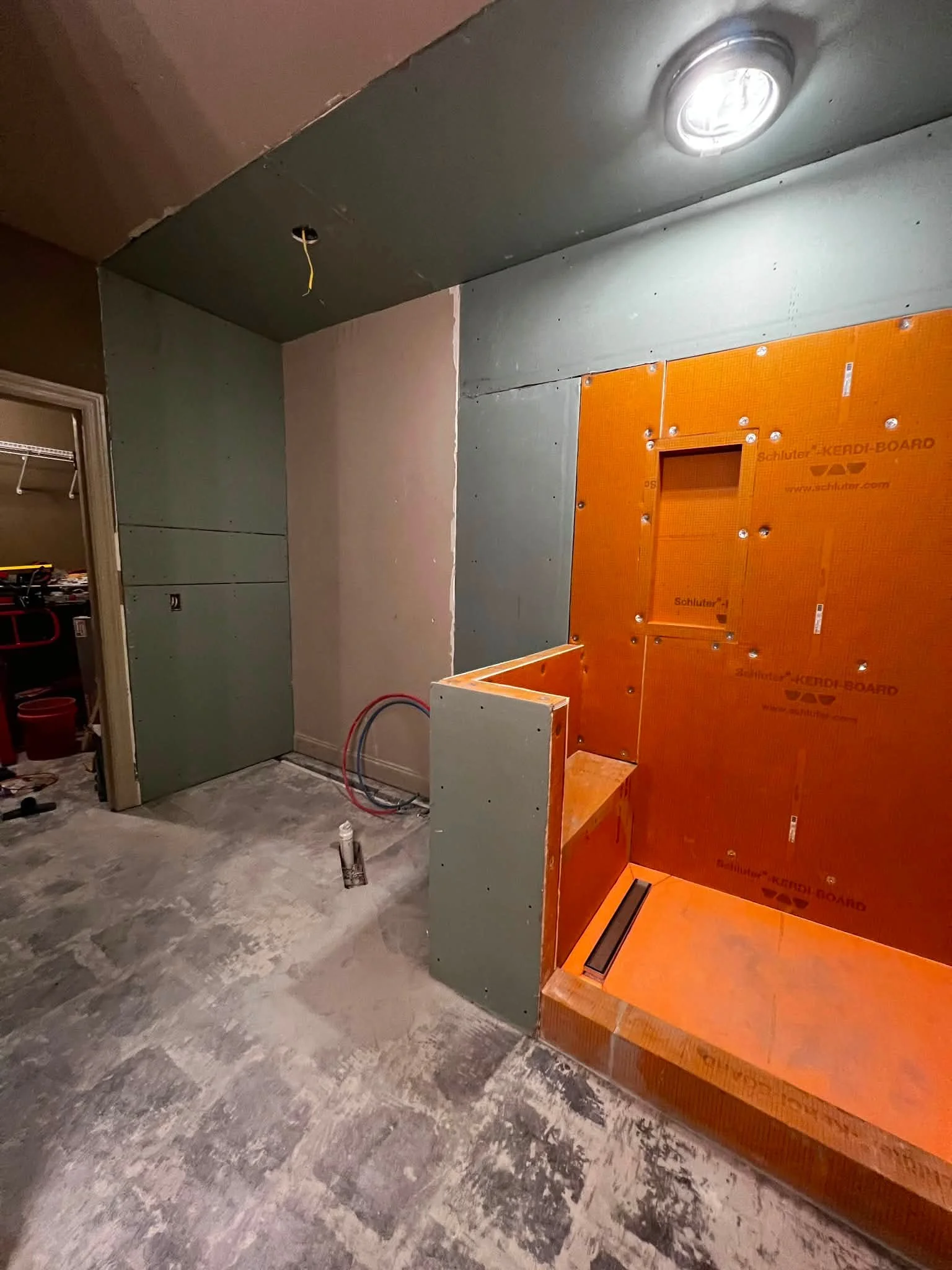 A room under construction with gray and orange drywall panels, an installed ceiling light, and exposed wiring. The floor is unfinished with a rough concrete surface.