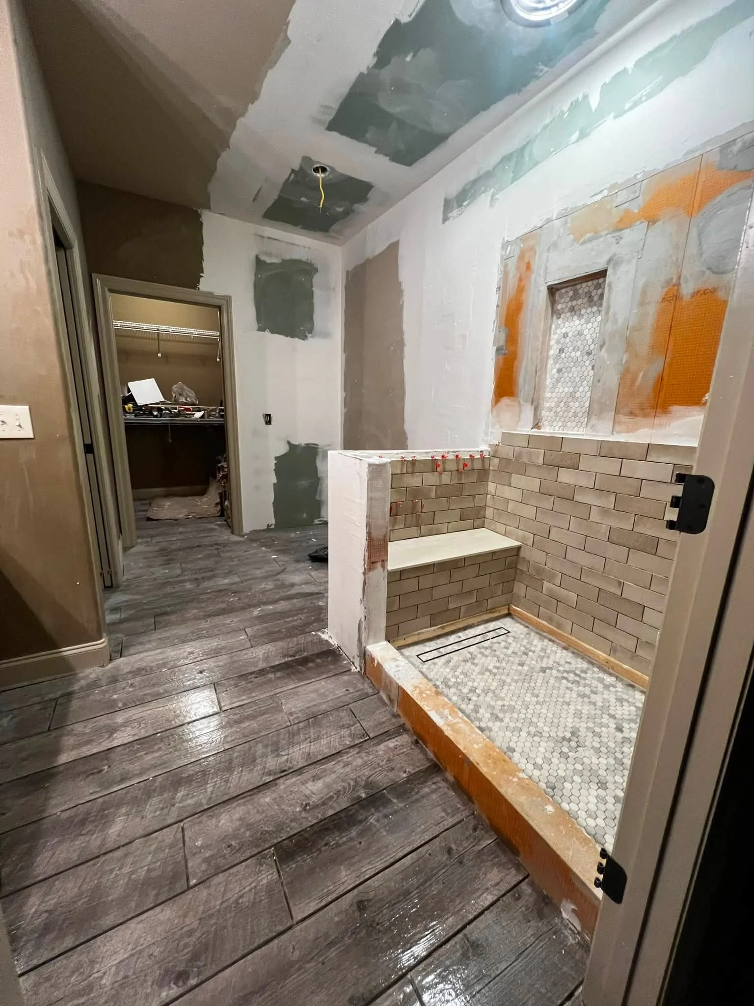 Interior bathroom under renovation with unfinished walls, a partially built shower area with brick and tile, and a doorway leading to a walk-in closet with cluttered shelves.