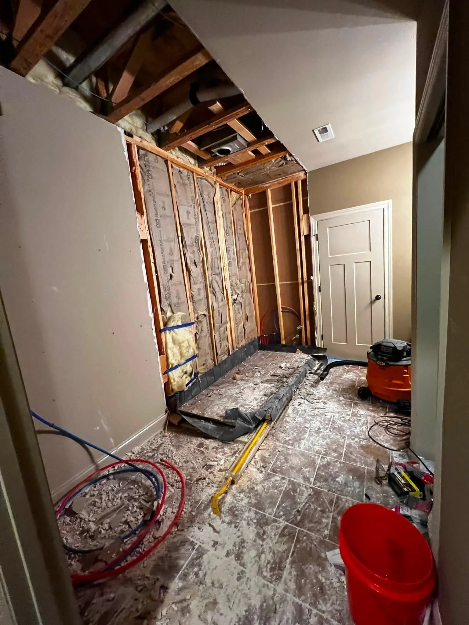 A room under renovation with exposed wall studs and insulation, construction tools, debris on the floor, and a white door.