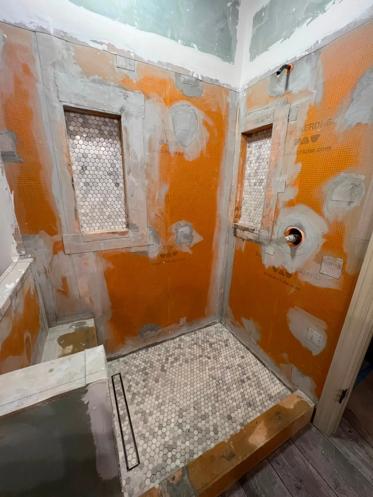 Bathroom under renovation with unfinished walls, orange waterproofing membrane, and a partially tiled floor. Pipes are visible on the wall, and protective tape covers window openings.