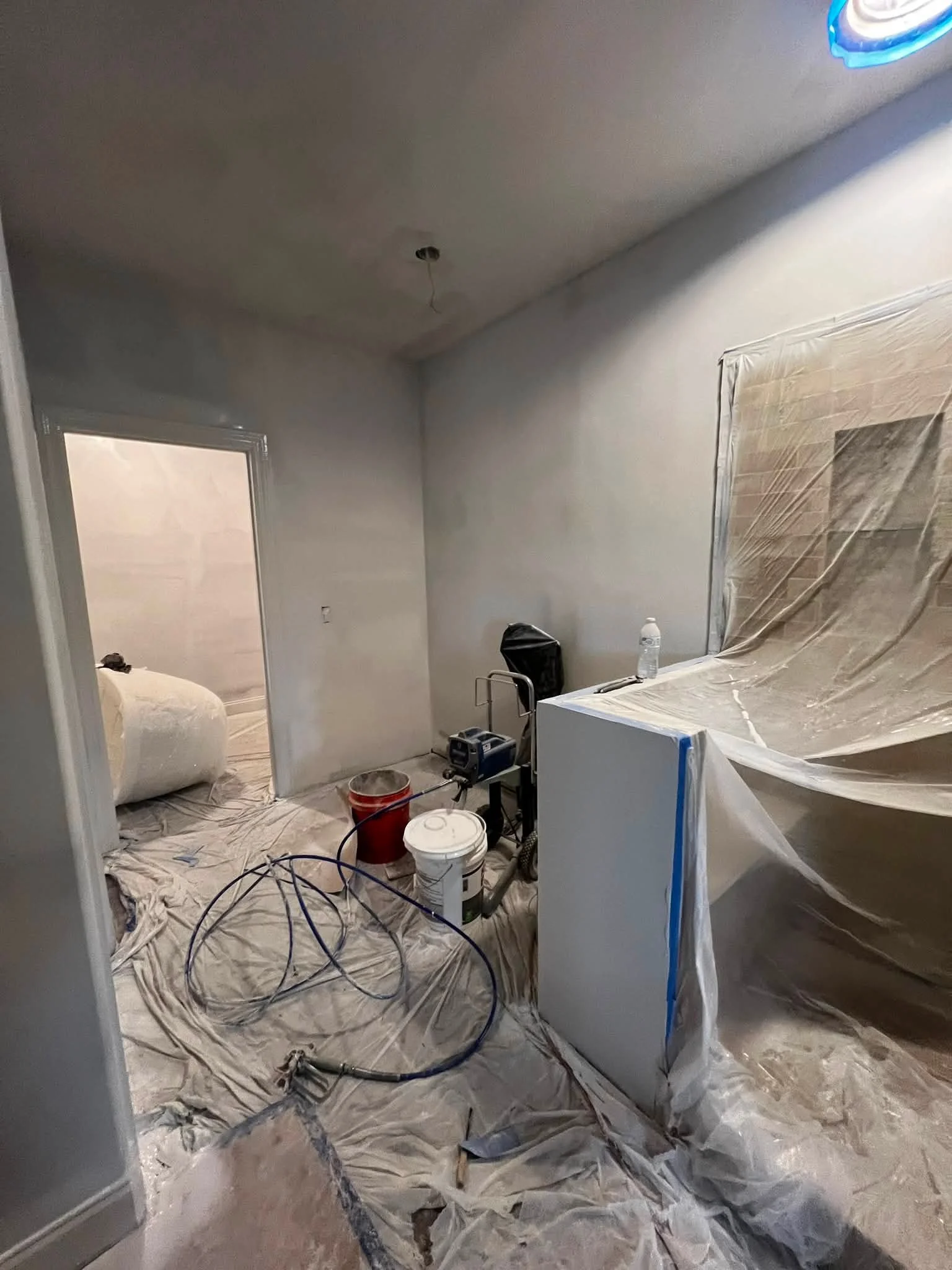 A room under renovation with plastic sheets covering the floor and furniture. There are tools, extension cords, buckets, and a water bottle visible. The ceiling appears to be in the process of being painted or redone.