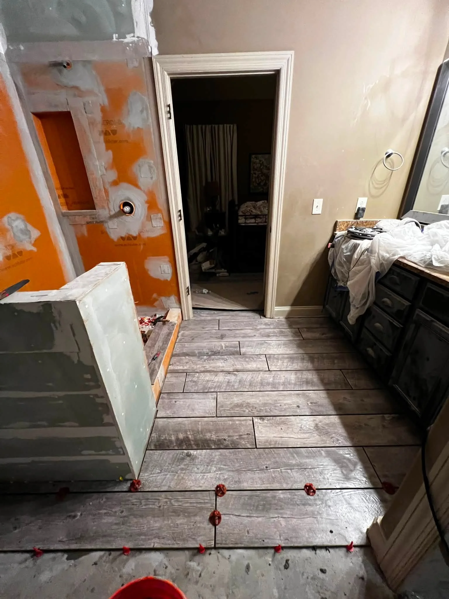 Partially renovated room with a doorway leading into a darker room, unfinished wall with drywall and wiring, a dresser on the right side covered with a white cloth, a mirror with ring hooks, and a floor with wood-like tiles. Construction materials an