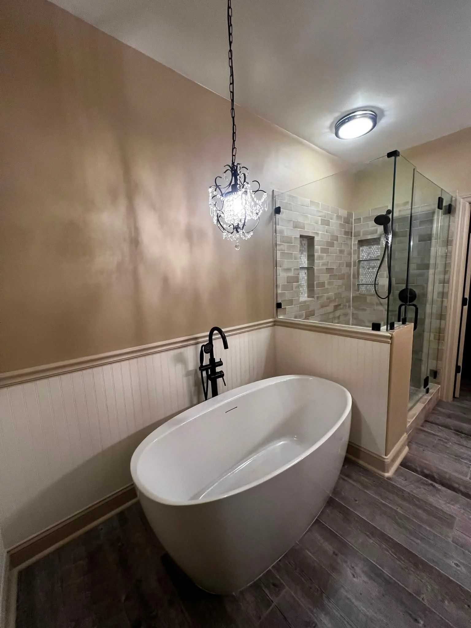 A bathroom with a freestanding white bathtub, a chandelier hanging from the ceiling, a glass-enclosed shower with brick-patterned tiles, a black towel hook, and dark wooden floorboards.
