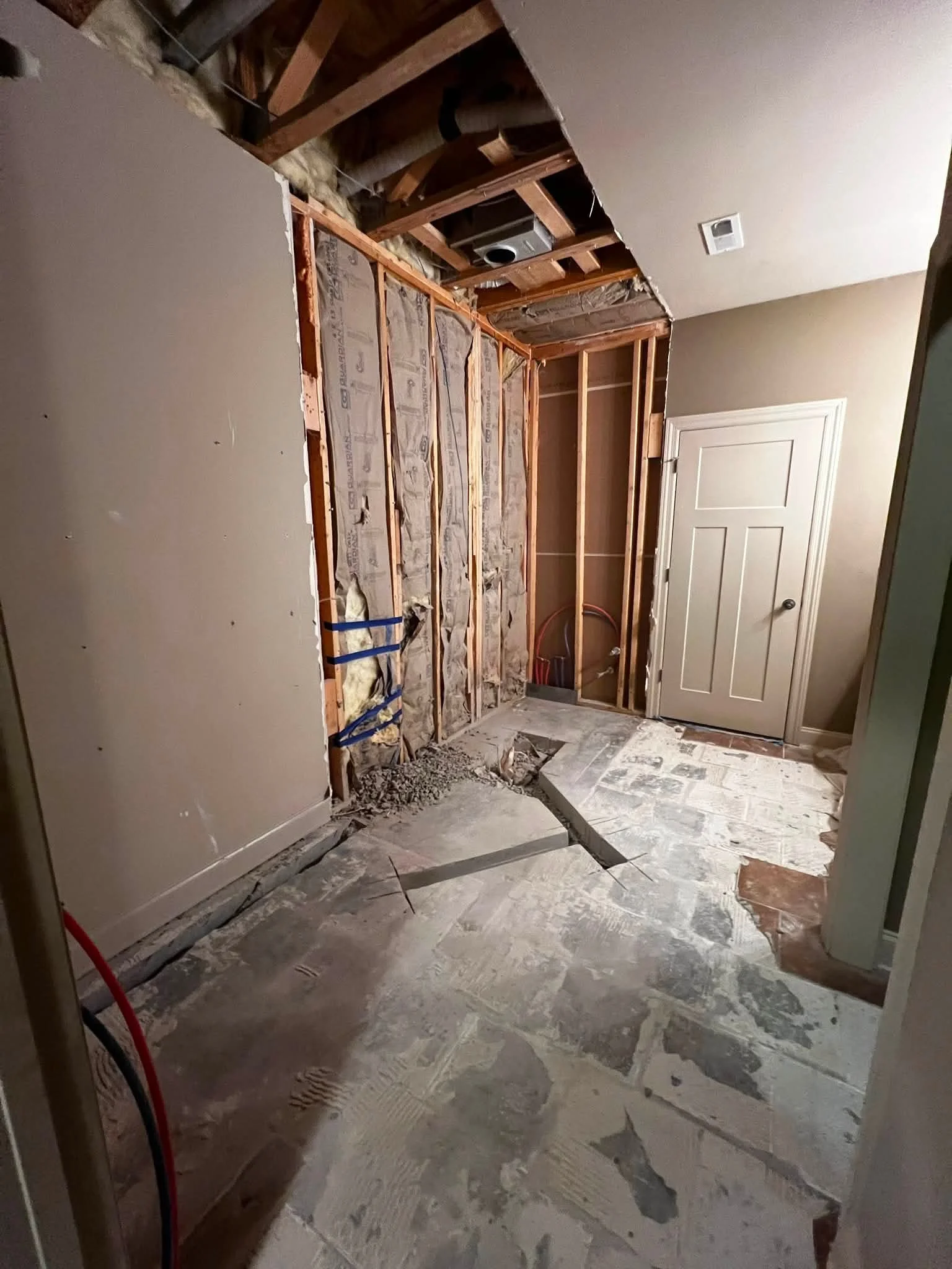 A room under construction with exposed wooden framing and insulation. The floor is partially tiled, and there is a closed door on the right side. Construction debris is visible on the floor.