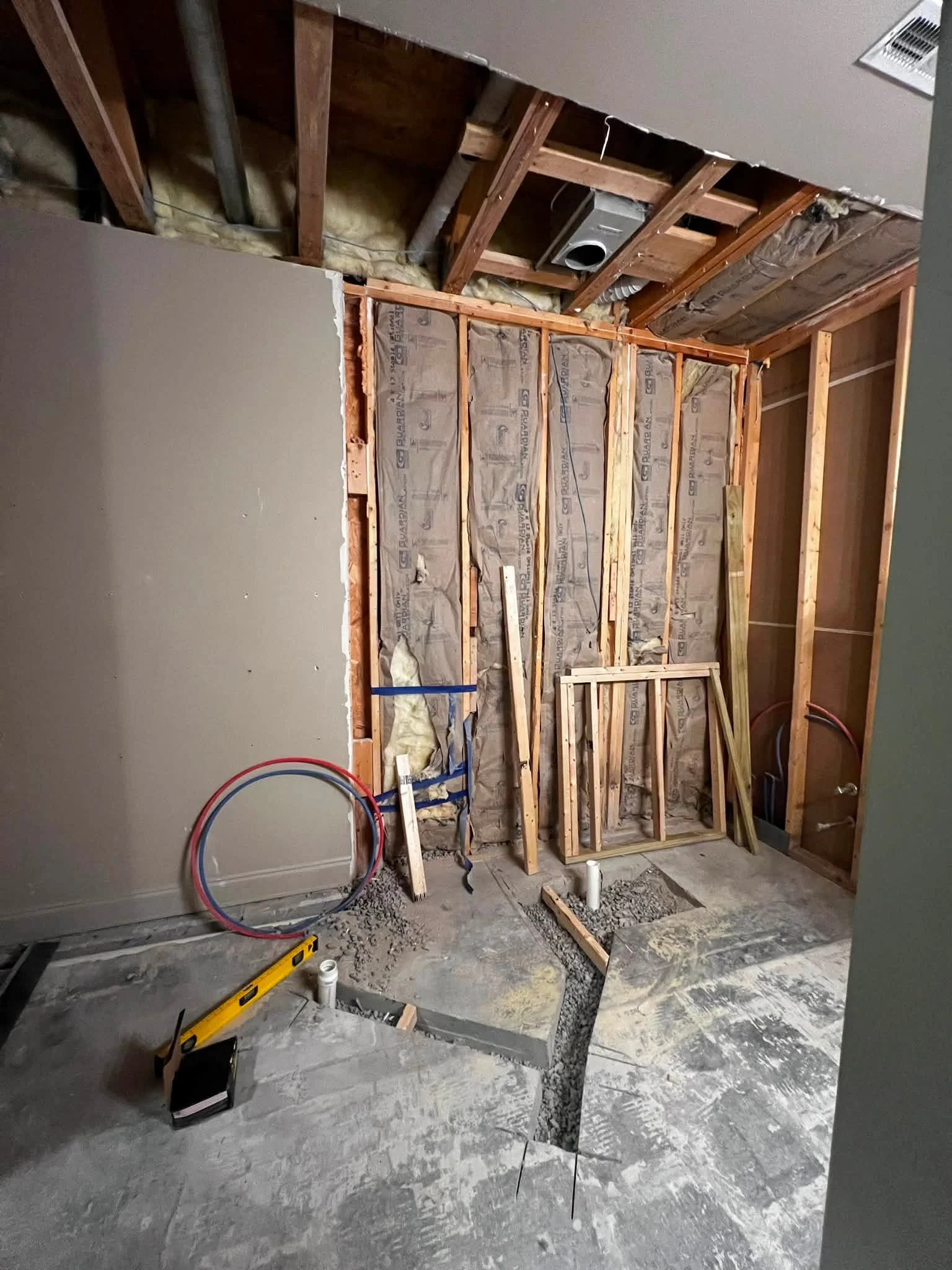 Interior of a room under construction with exposed wooden studs, insulation, and plumbing. Construction tools and materials are on the unfinished concrete floor.