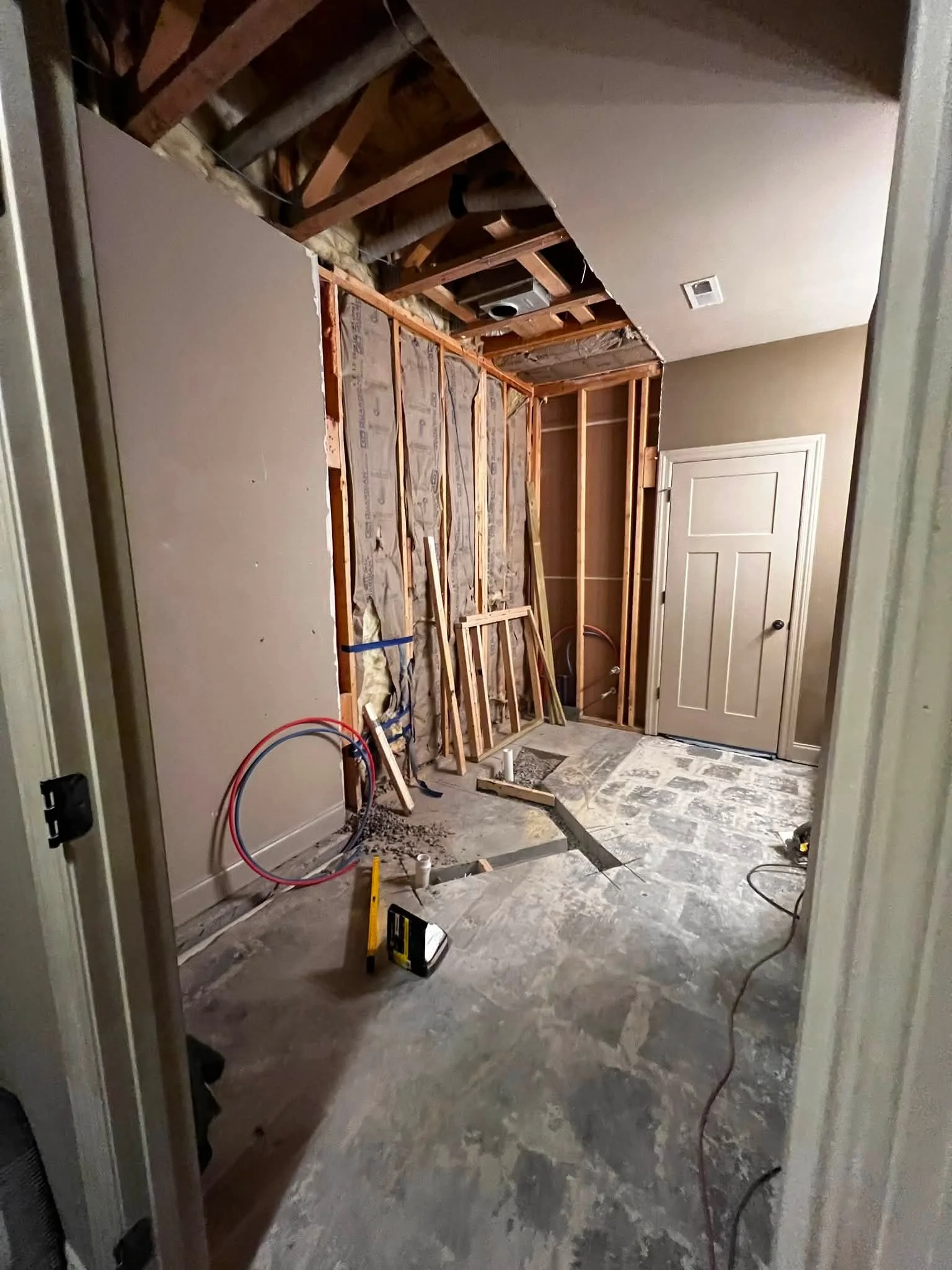 Room under renovation with exposed wall framing, insulation, and partially removed drywall. Construction tools and materials are on the floor, and a door is closed on the right side.