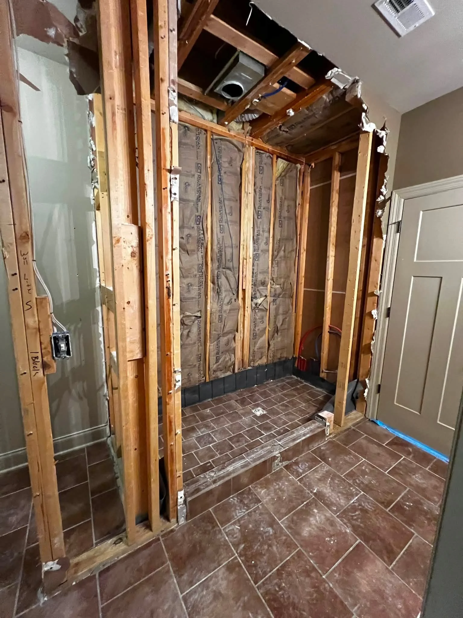 Partially demolished bathroom with exposed wall studs, insulation, and plumbing, with brown tile flooring and a closed door.