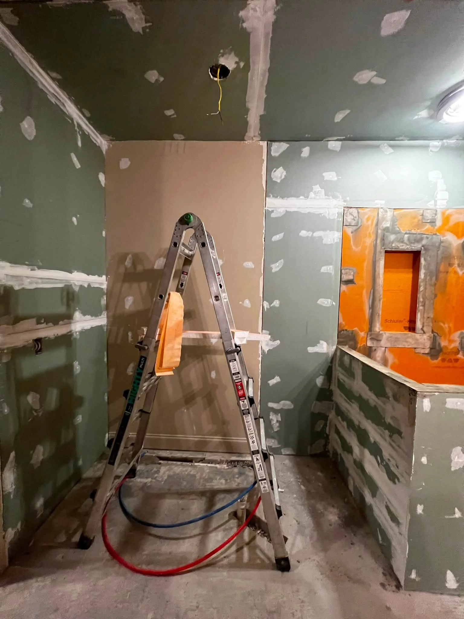 Interior of a room under construction or renovation with drywall partially installed and mudded. A step ladder is in the center with red and blue hoses or cords on the floor. The ceiling has an electrical box with wiring hanging down. The walls have 