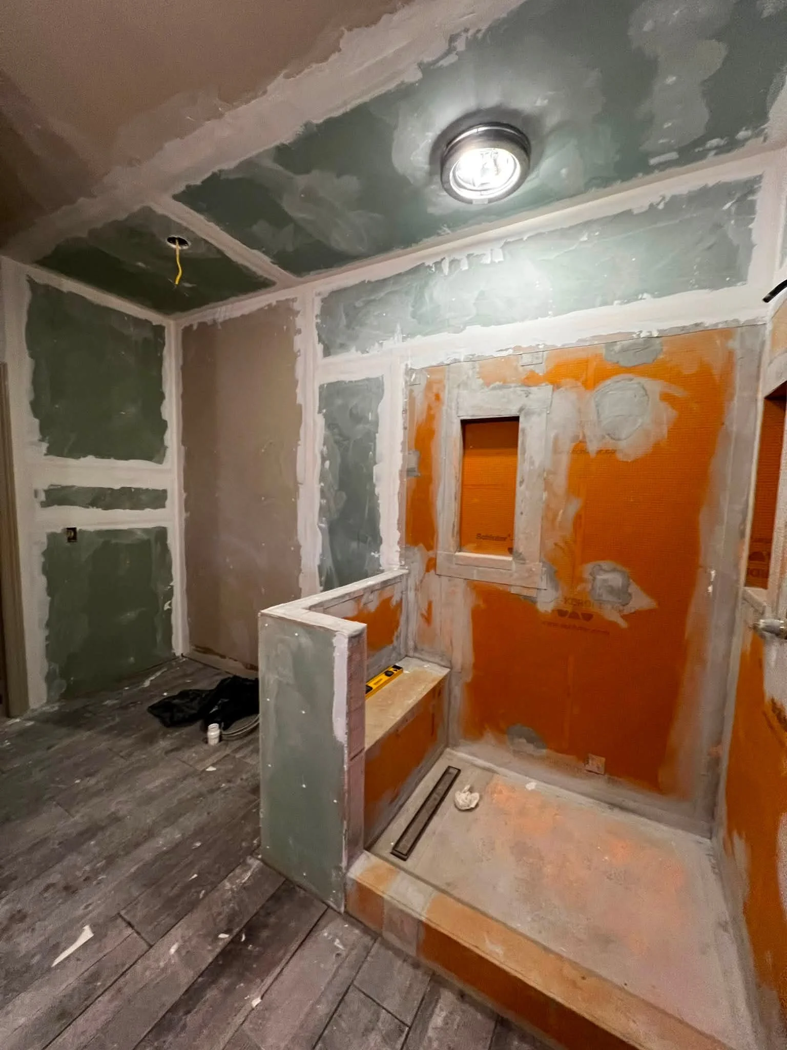 A partially finished room with drywall on the walls and ceiling, an unfinished floor with wooden planks, a circular ceiling light fixture, and an opening in the wall with orange insulation behind it.
