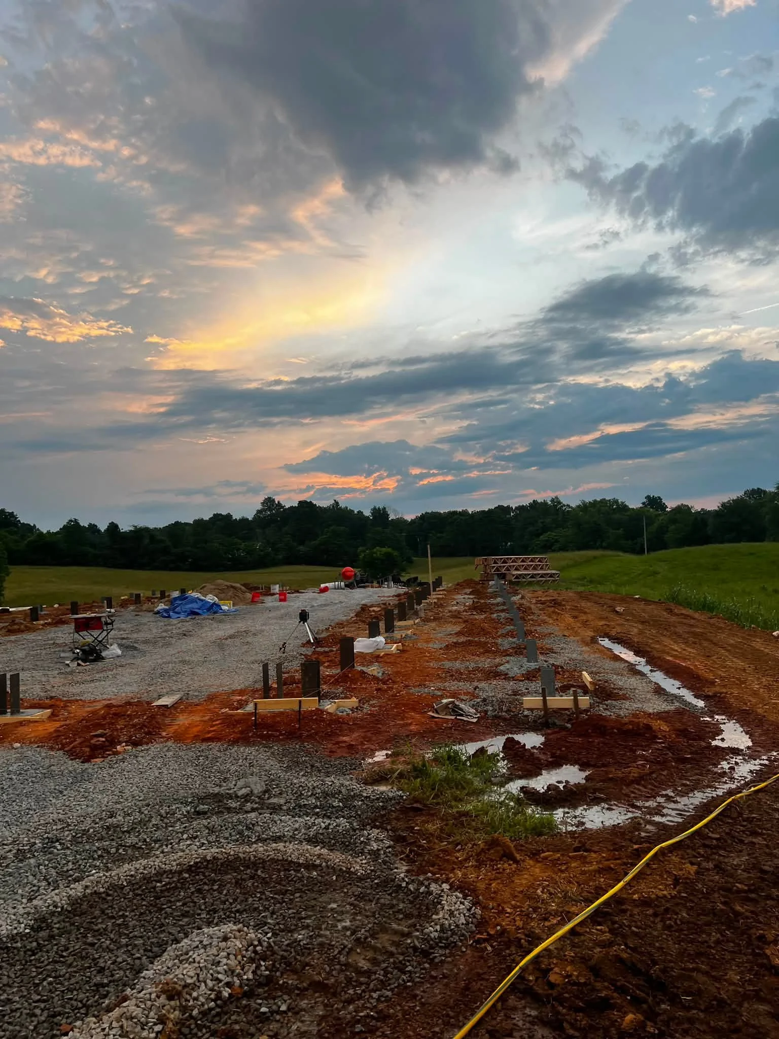 A construction site at sunset with a dirt and gravel road, construction materials, and equipment, set against a backdrop of green fields, trees, and a partly cloudy sky with sunlight streaming through.