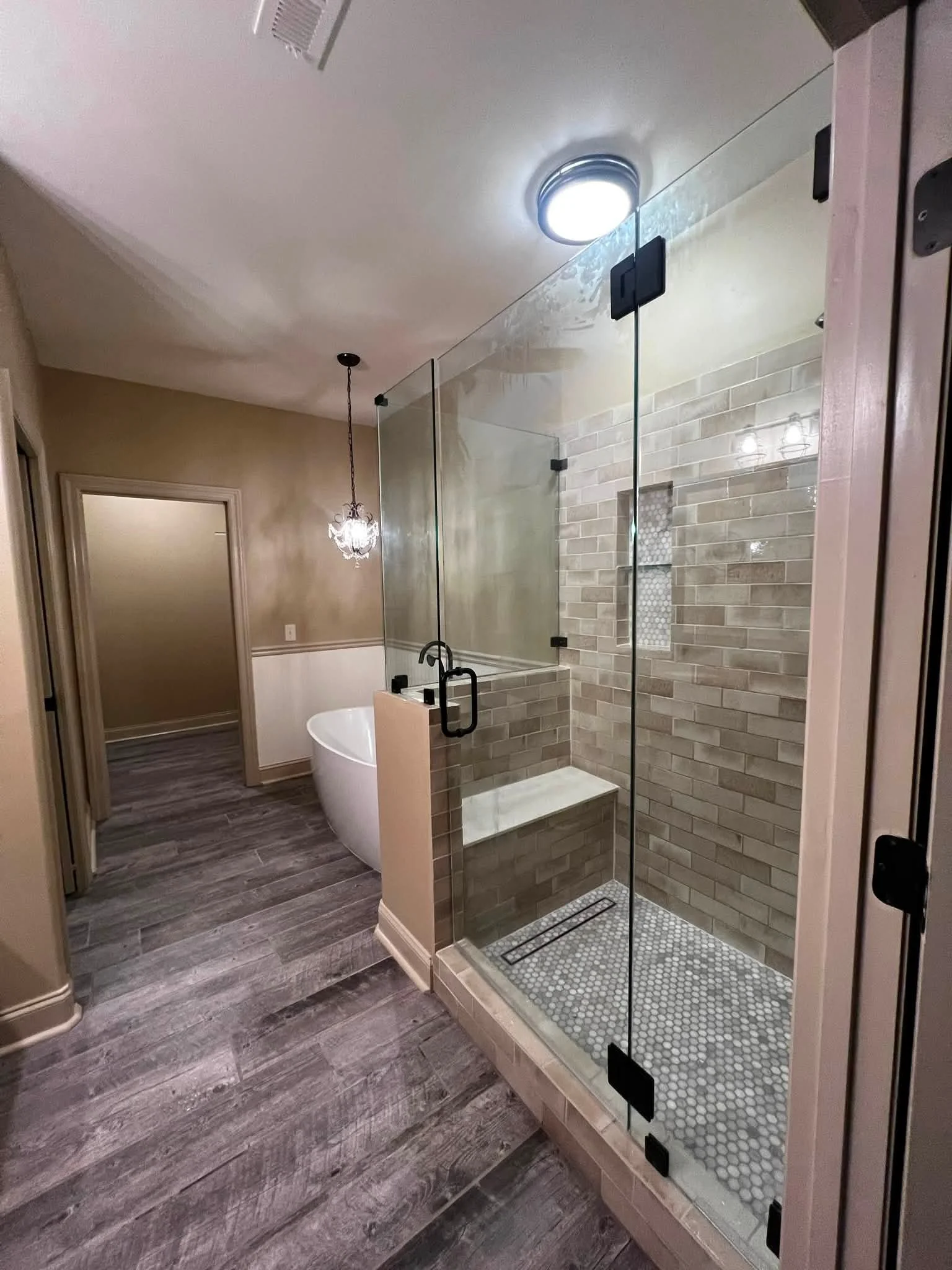 A modern bathroom with a glass-enclosed shower area featuring beige subway tiles and small round tile flooring, a freestanding bathtub, wooden flooring, and a chandelier. The ceiling has a round light fixture and an air vent.