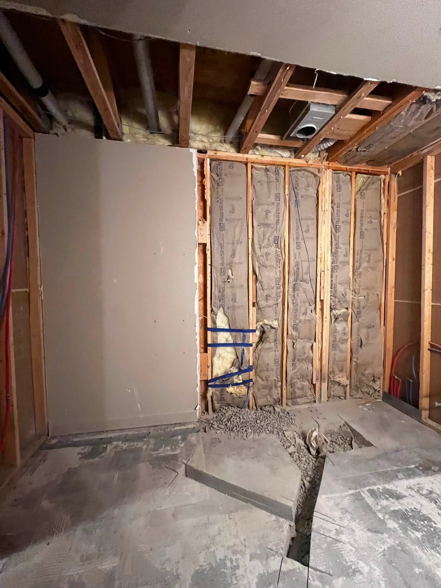 Interior of a room under renovation with exposed wall studs and insulation, a partially finished drywall on the left, and construction debris on the floor.
