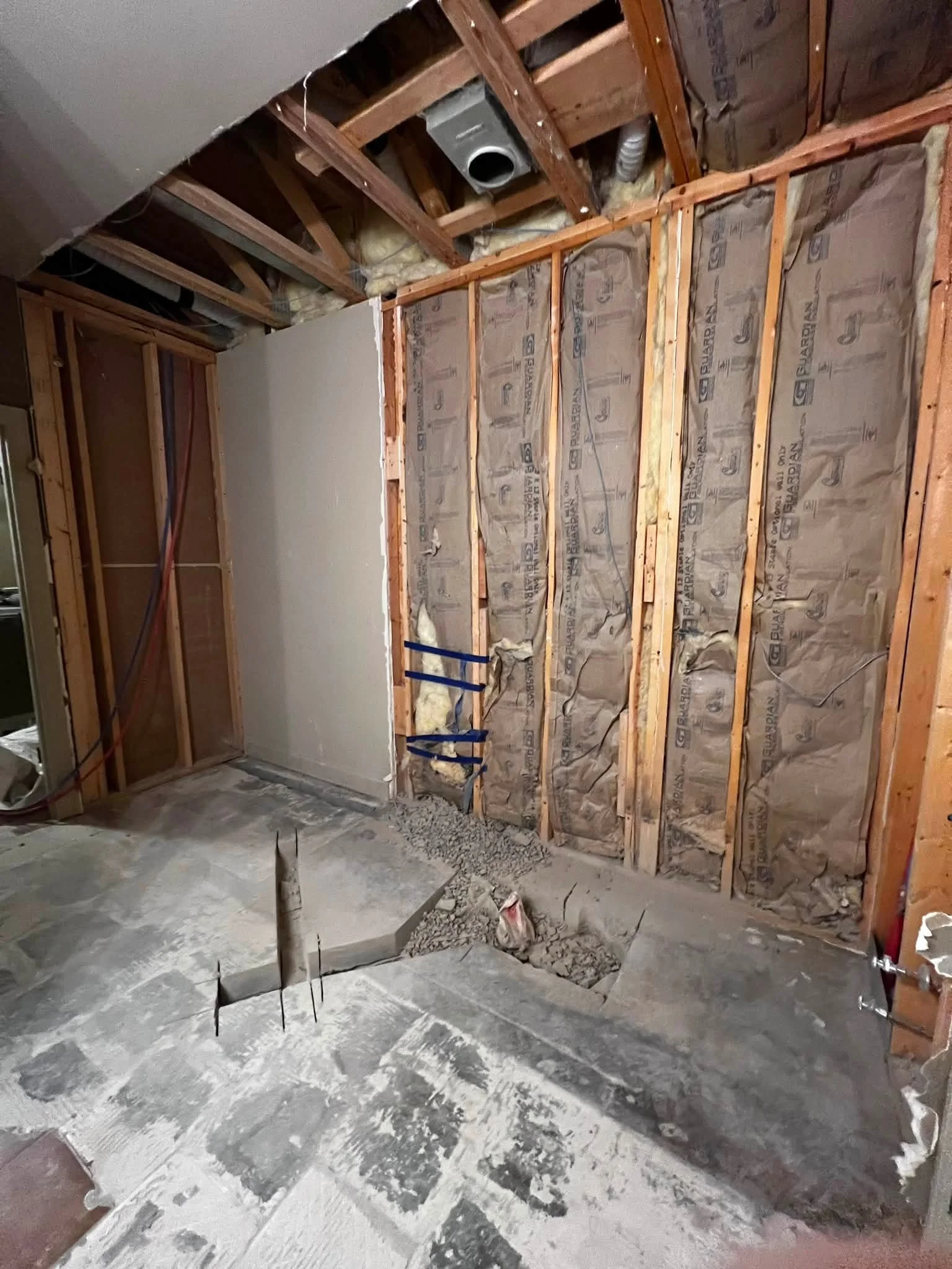 Interior of a room under construction with exposed wooden framing, insulation, and a concrete floor. There are some construction tools and materials visible, including a large hole in the floor.