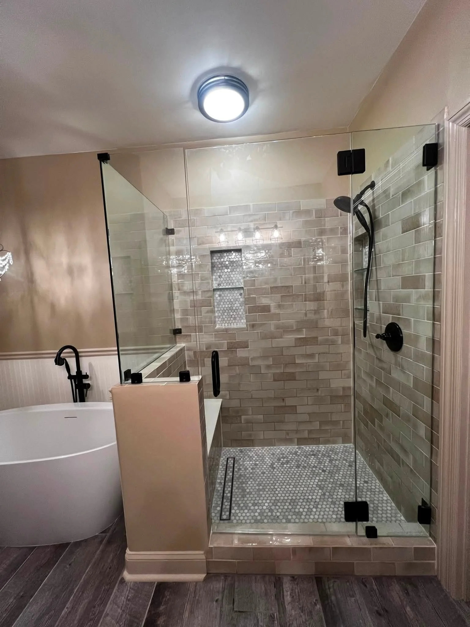 Modern bathroom with a glass shower enclosure, beige and gray tiled walls, a black showerhead and fixtures, a freestanding white bathtub, and a white shower seat, with a dark wooden floor and ceiling lighting.