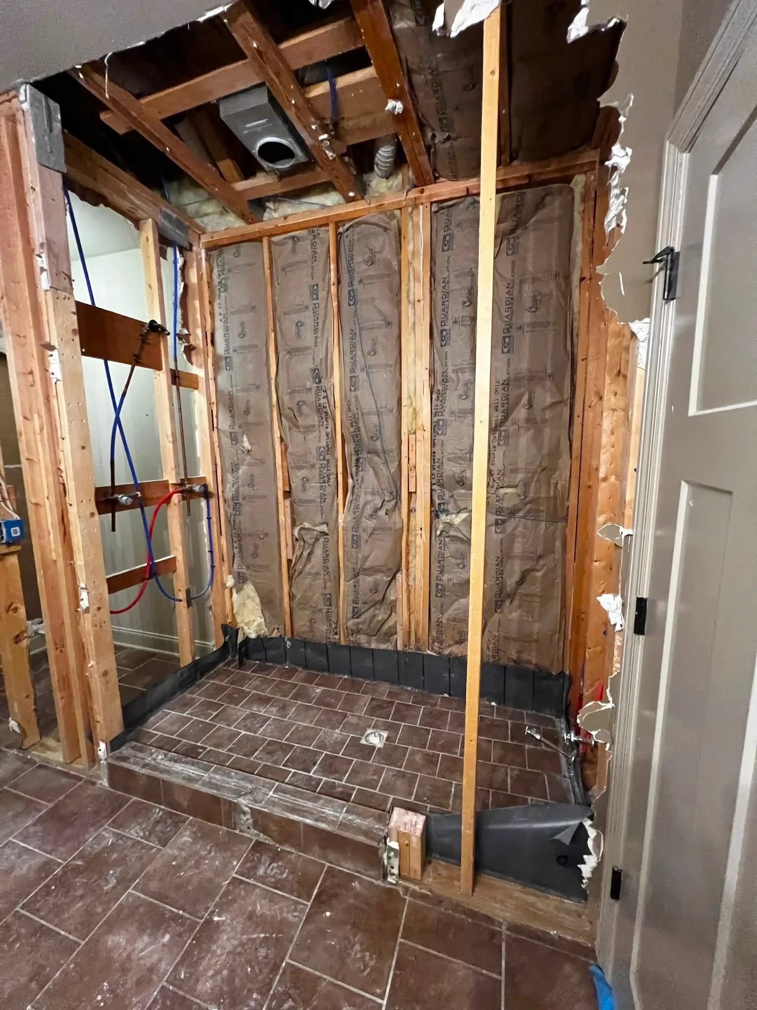 Interior of a room with wall removed, exposing wooden framing and insulation for a bathroom renovation. The floor has dark brown tiles, and there are pipes and ducts visible within the wall cavity.