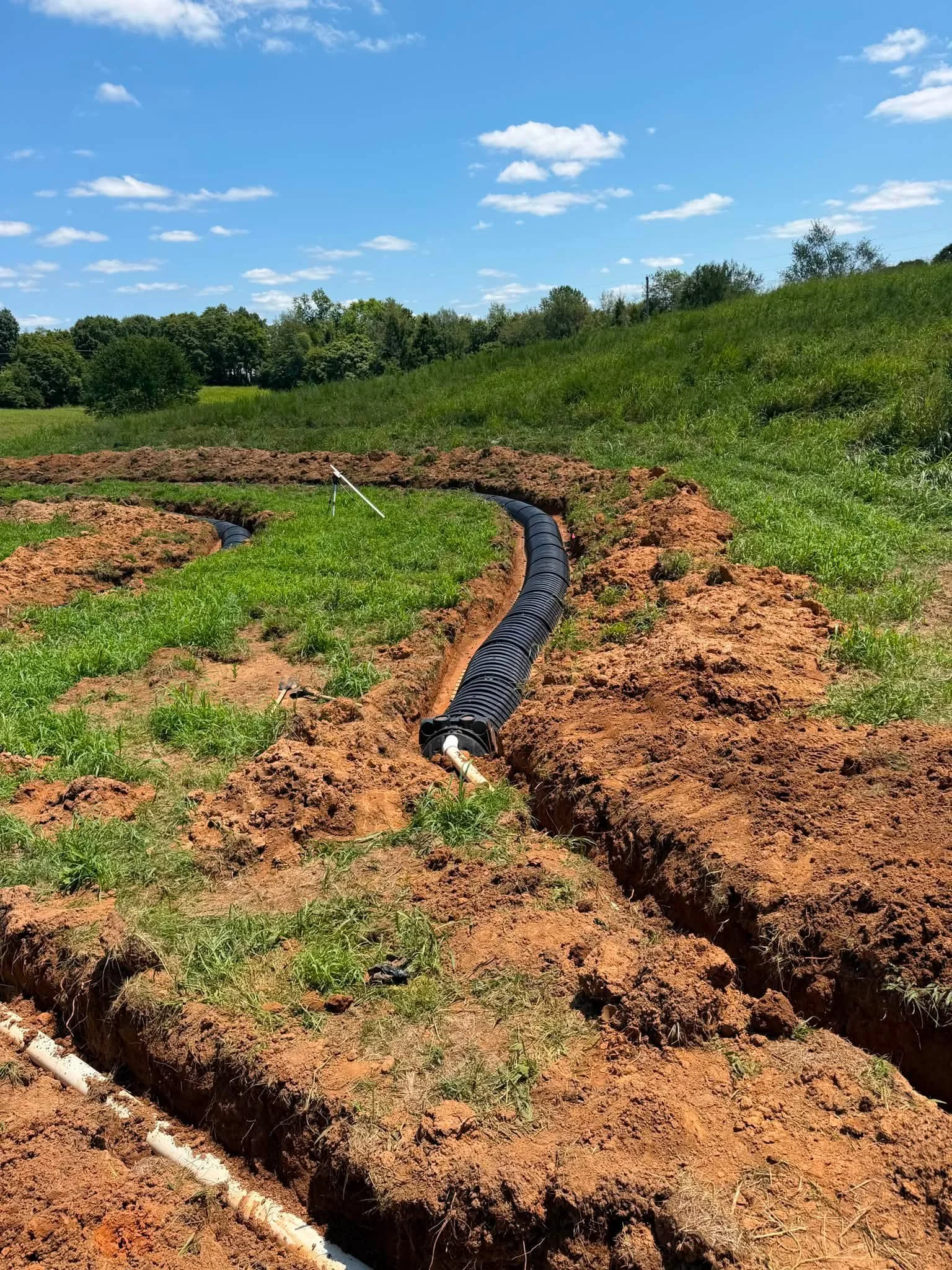 A black corrugated pipe installed in a trench in a grassy field, with dirt mounded on either side, under a partly cloudy blue sky.