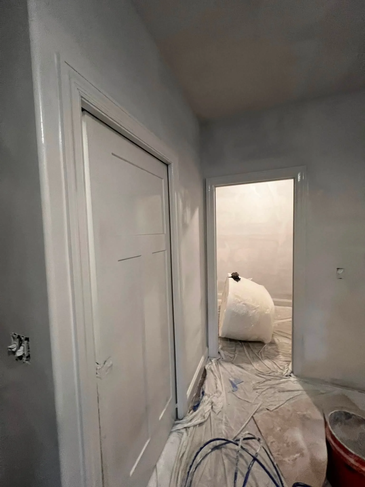 A hallway under construction or renovation with plastic sheeting on the floor, paint tools, and a room visible through an open doorway. The walls are painted light gray.