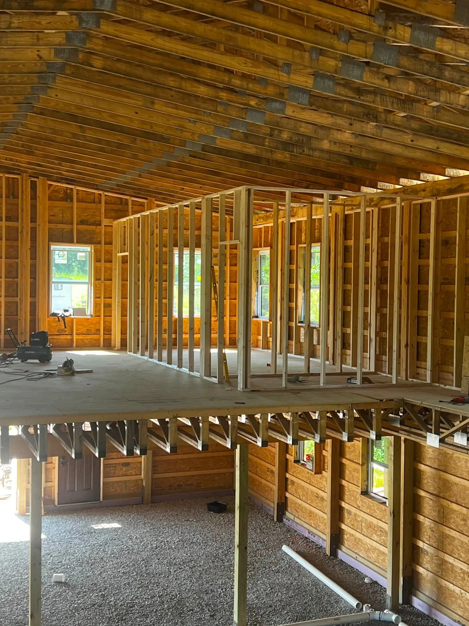 The interior of a house under construction showing wooden framing, with exposed walls, ceiling, and windows. There are construction tools and materials on the floor.