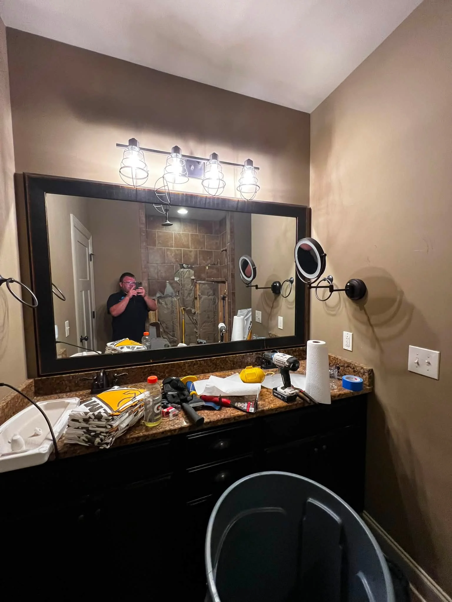 A bathroom vanity mirror with overhead lights, a cluttered granite countertop with various items including paper towels, a drill, and other small tools, and a person taking a selfie in the reflection. The room features beige walls and a shower with a
