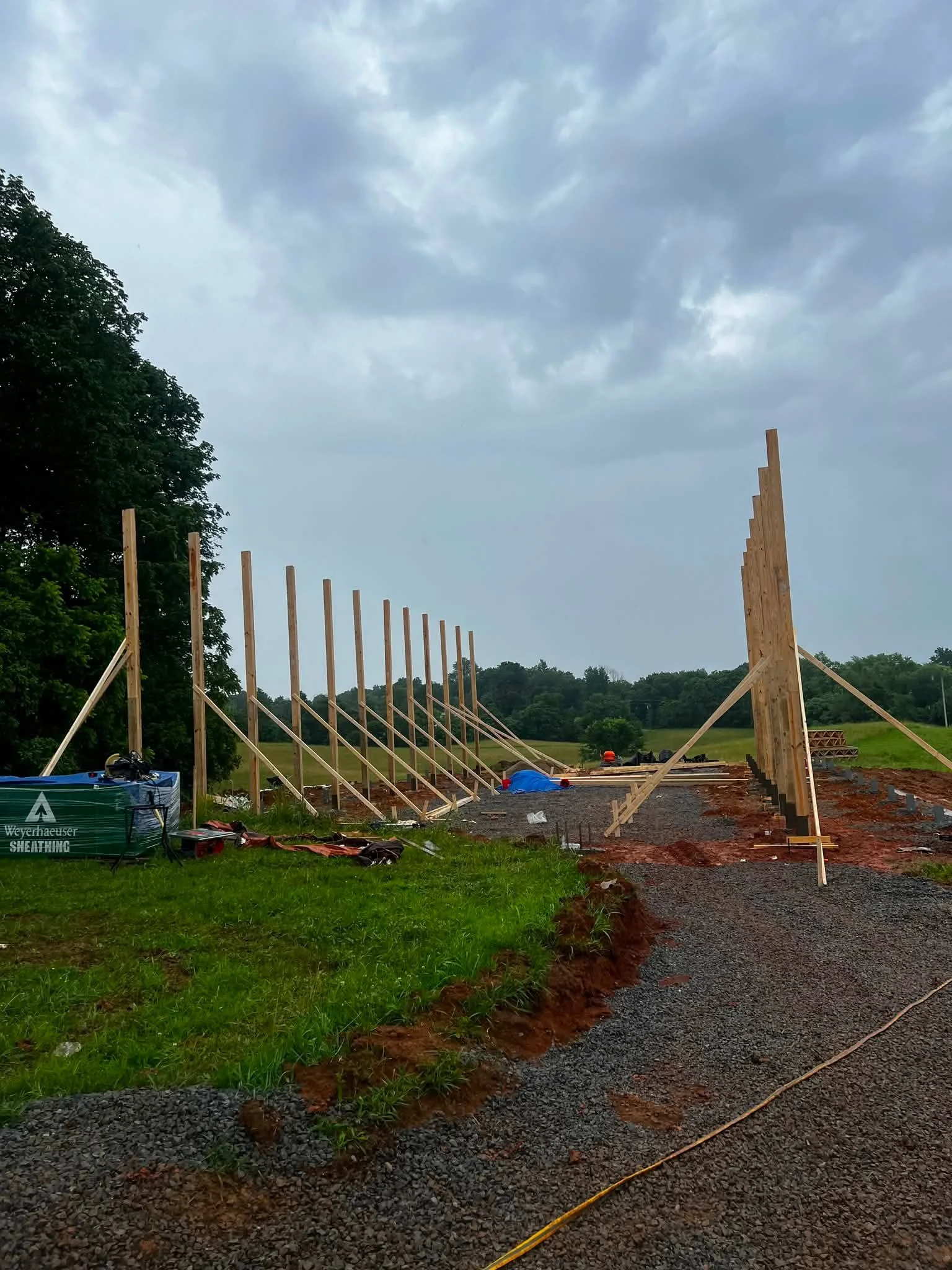 Construction site with wooden posts, gravel pathway, and overcast sky, surrounded by greenery.