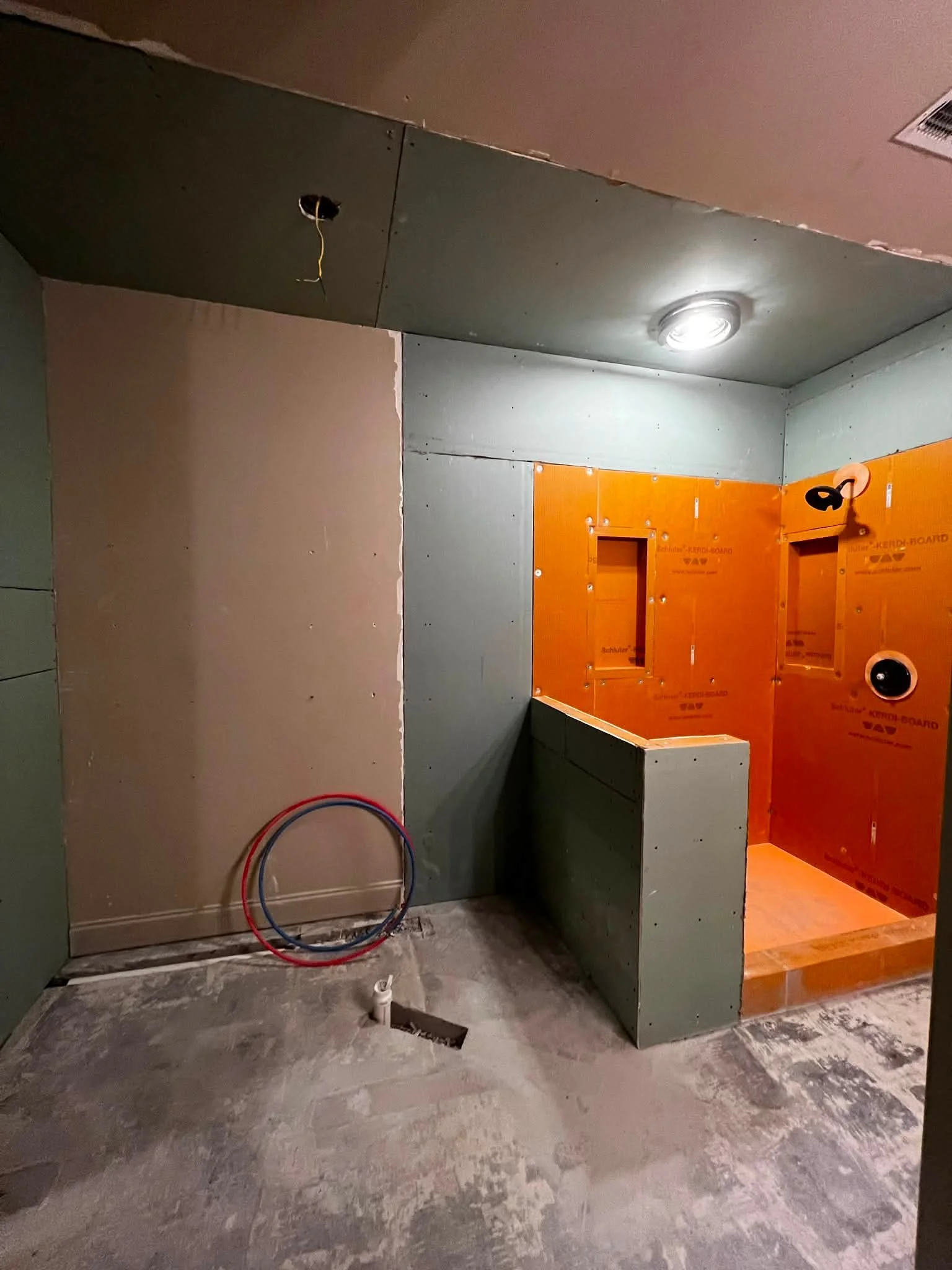 Construction site with unfinished drywall and orange KERDI-BOARD walls, empty open spaces, exposed electrical wiring, and rough concrete flooring.