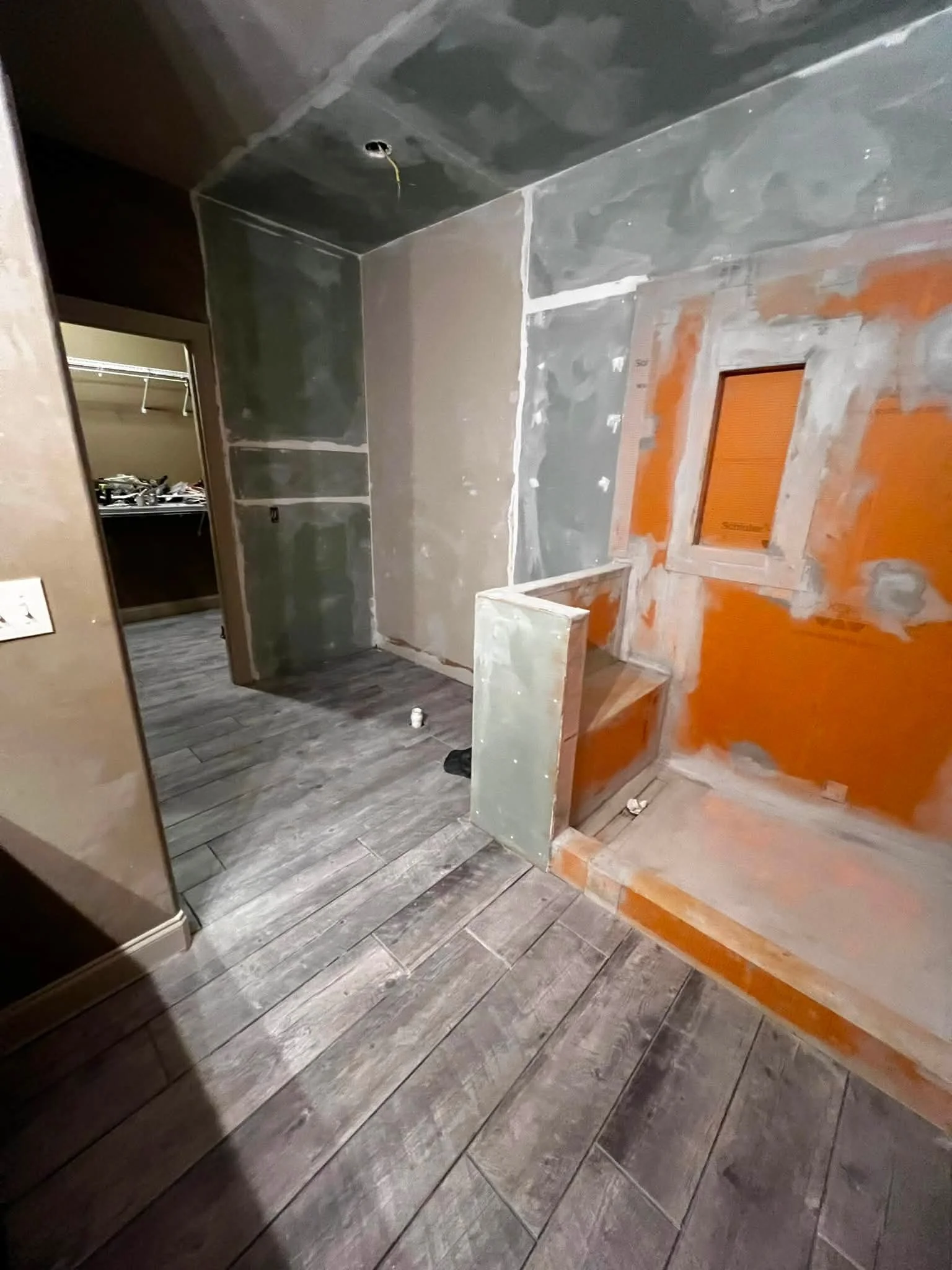 A partially constructed room with drywall installation in progress. The floor is covered with wood-look tiles. There are cut-out spaces in the drywall, possibly for windows or vents, and an open closet with hanging rods is visible in the background.