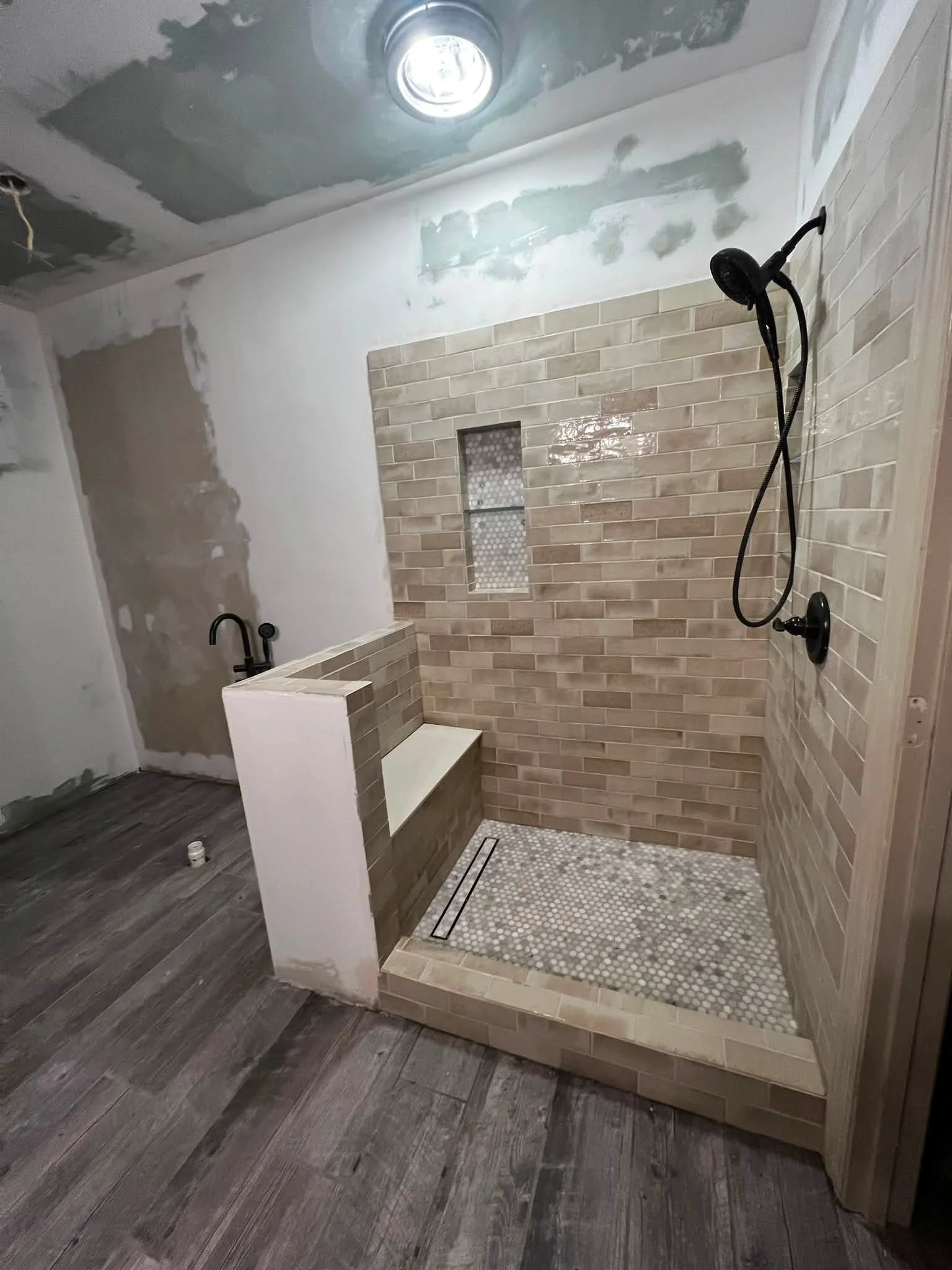 Bathroom shower area under renovation with tiled wall, built-in bench, and showerhead, with unfinished ceiling and flooring.