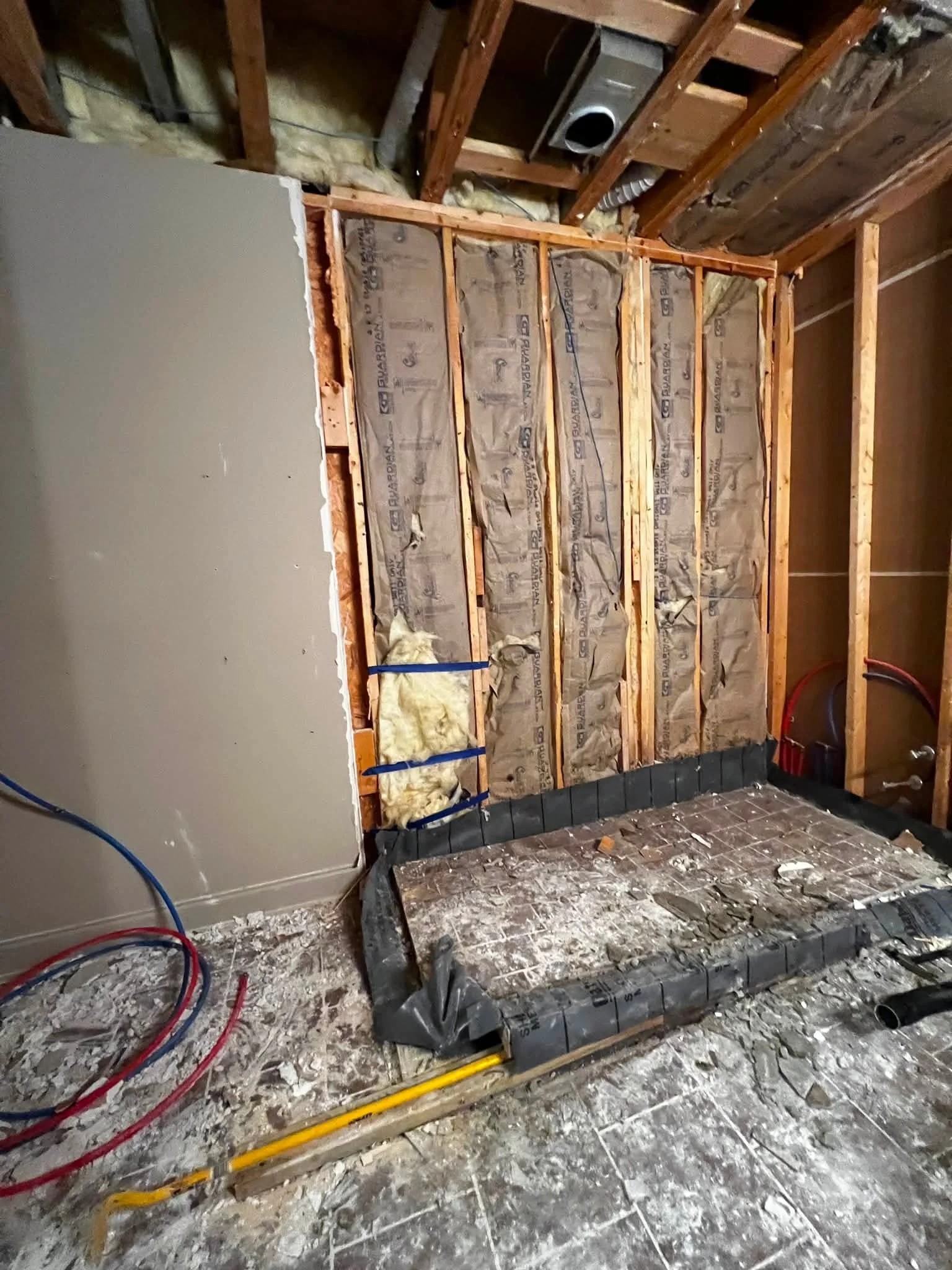 Interior of a room under construction with exposed wooden framing, insulation, electrical wiring, and a partially installed or removed appliance or fixture. Debris and construction materials are scattered on the floor.