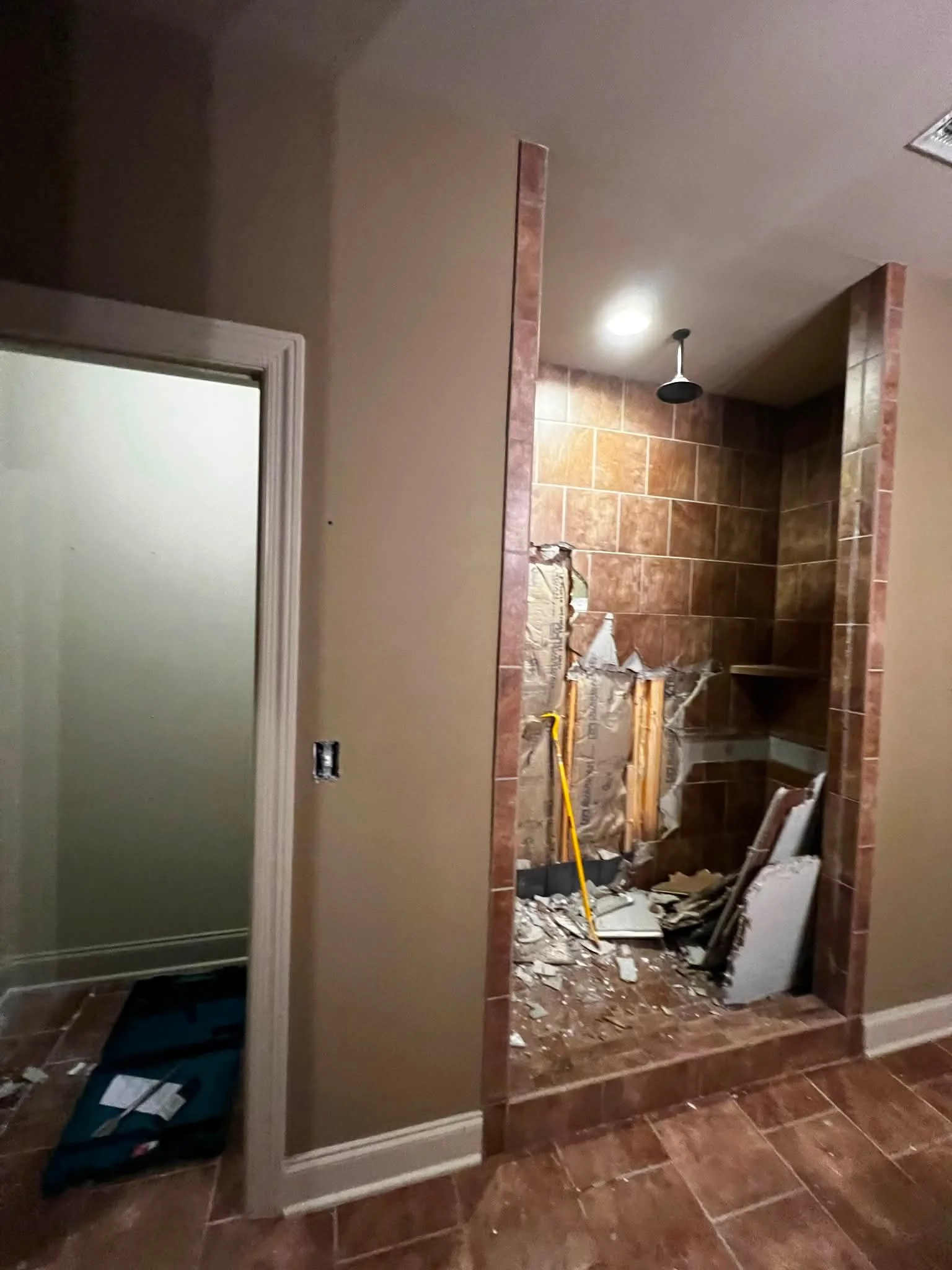 Bathroom with a shower area under construction, tools, debris, and partially installed tiles, with a beige wall and a white door frame in the foreground.