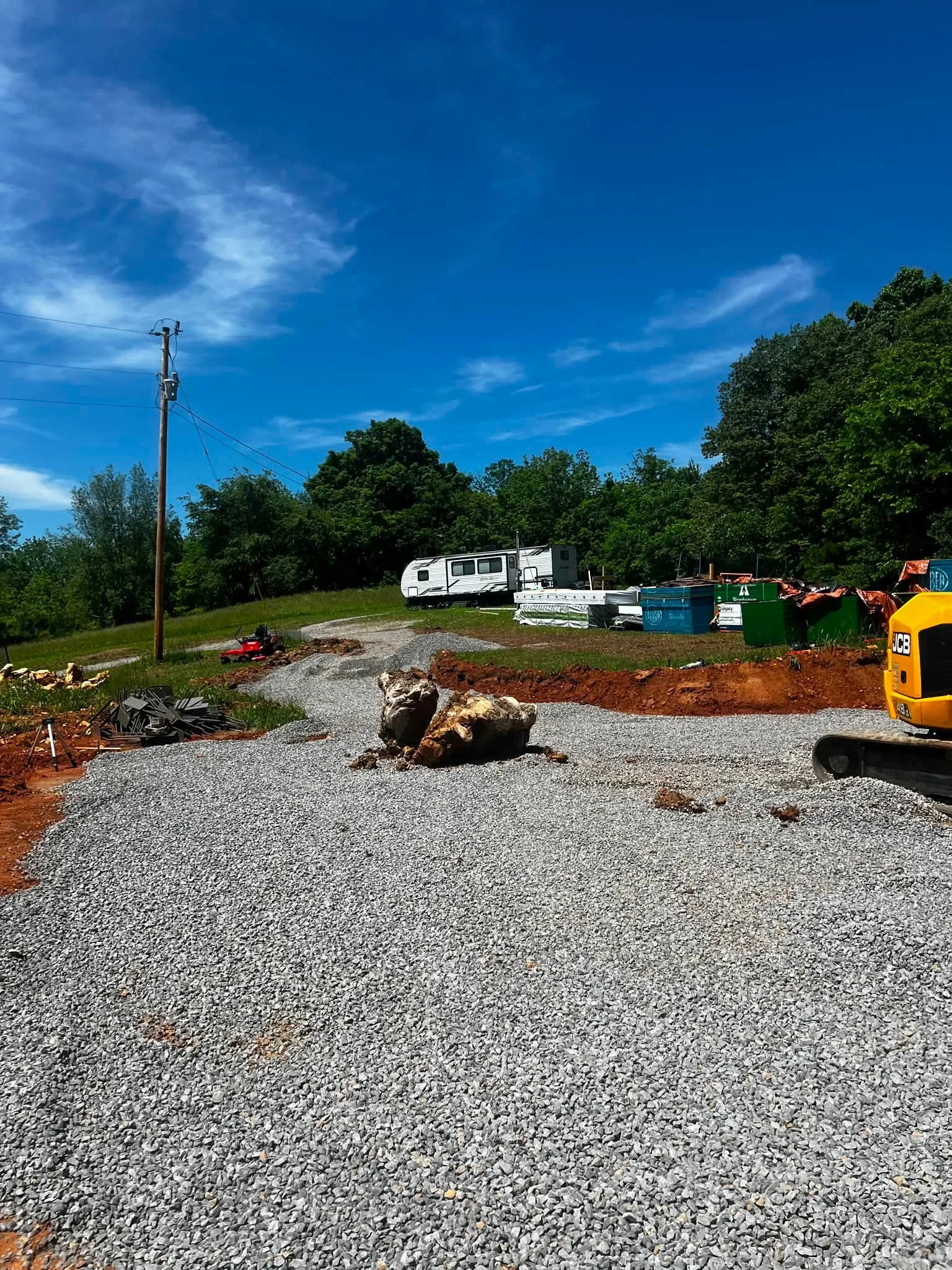 A gravel road with large rocks, construction equipment, a row of green and blue dumpsters, a small white camper trailer, trees in the background, and a bright blue sky with scattered clouds.