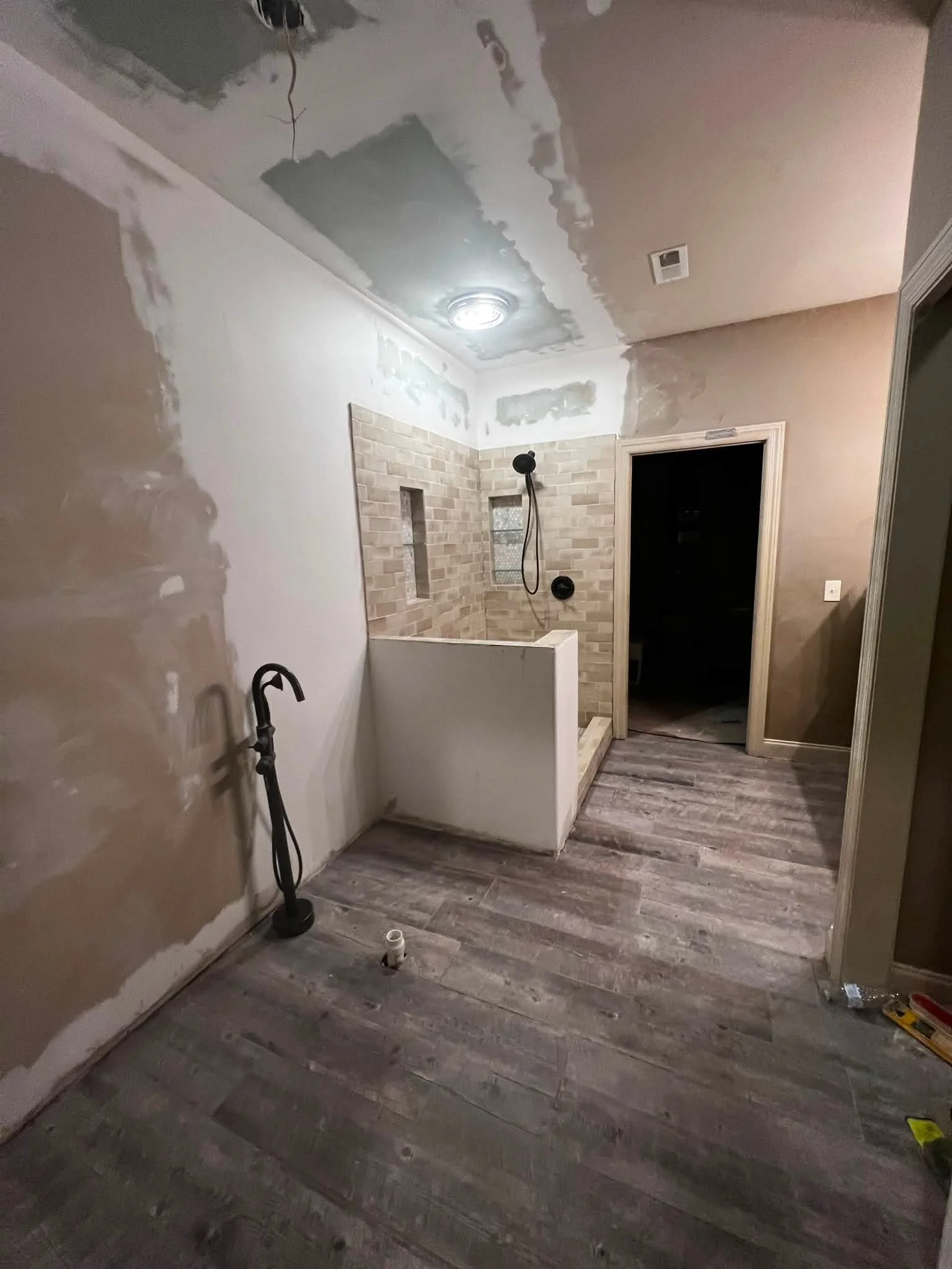 Bathroom under renovation with partially finished walls, a shower area with brick-patterned tiles, a ceiling light, and flooring with wood-like planks.