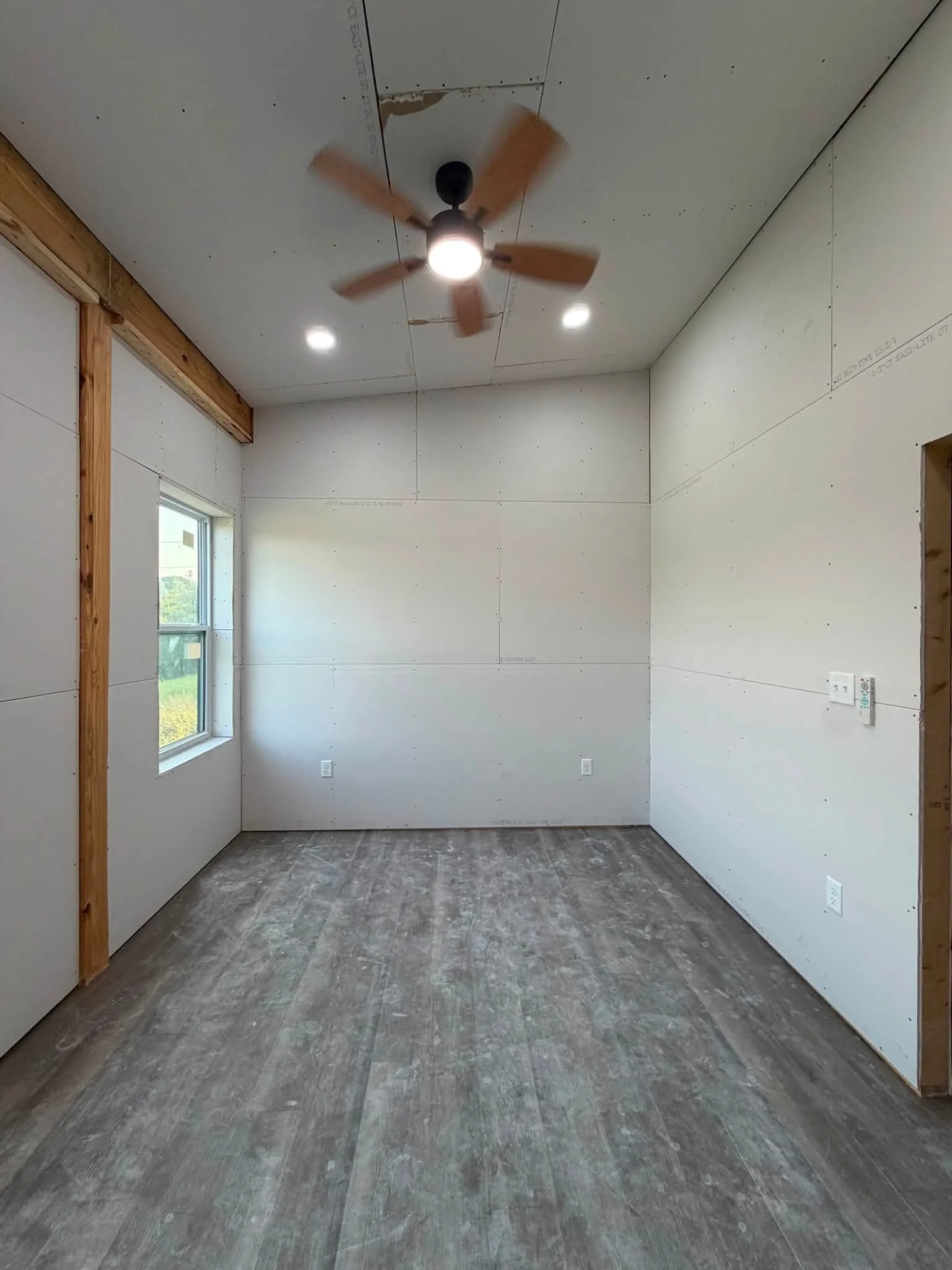 An unfinished room with drywall walls and ceiling, a ceiling fan, a window, and electrical outlets.