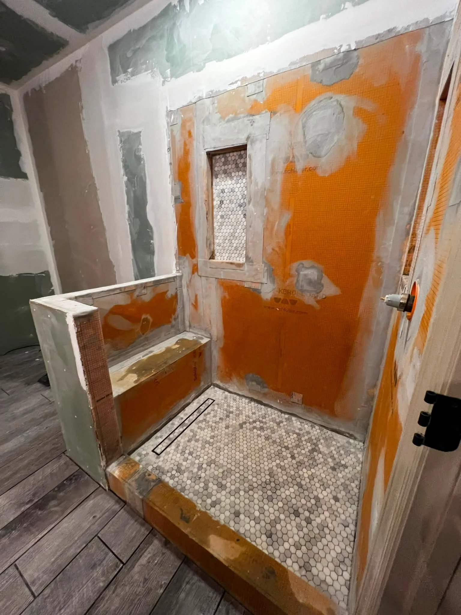 Bathroom under renovation with orange waterproofing membrane, partially installed tiles, and a shower drain on a wooden floor.