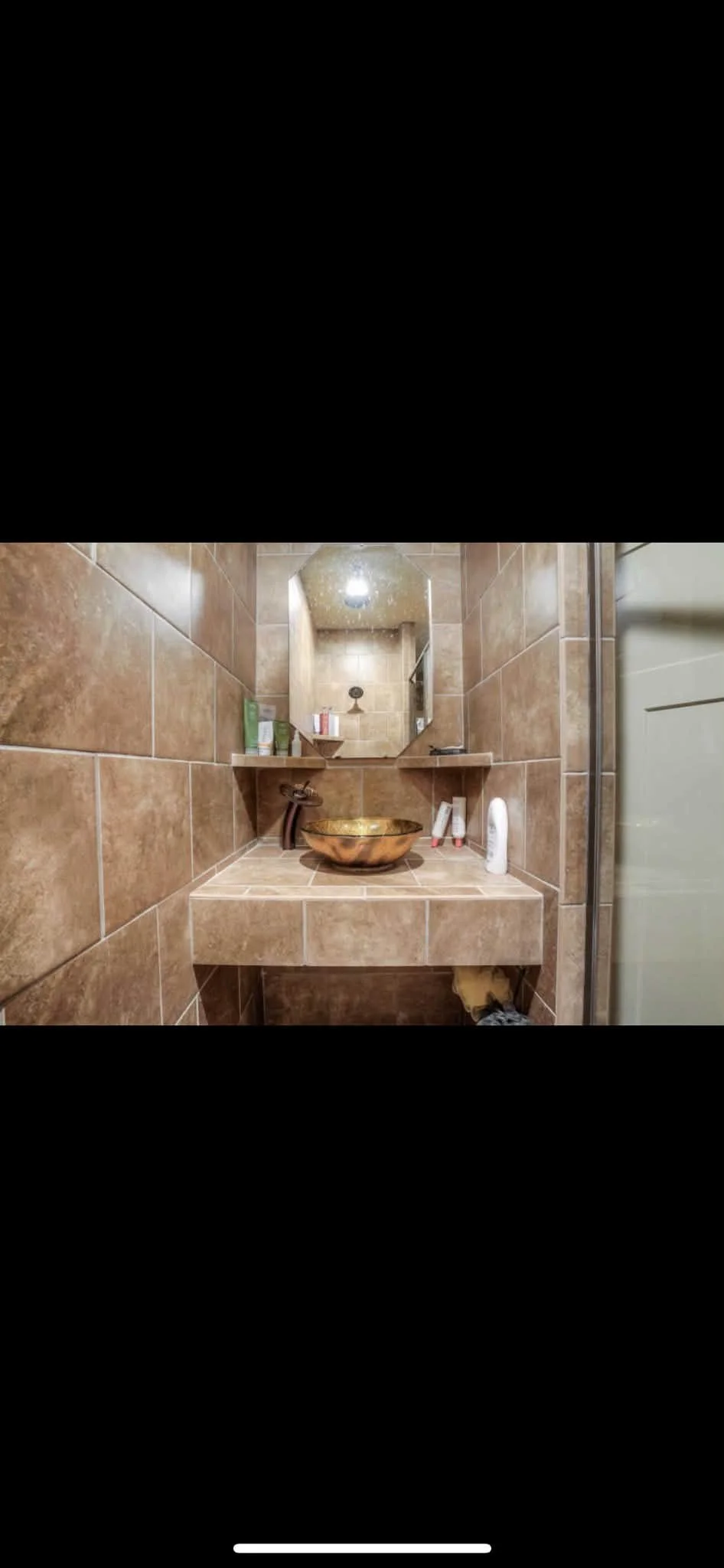 Bathroom with brown tiled walls, a mirror, a sink with a copper bowl, toiletries on shelves and counter, and partially visible shower door.