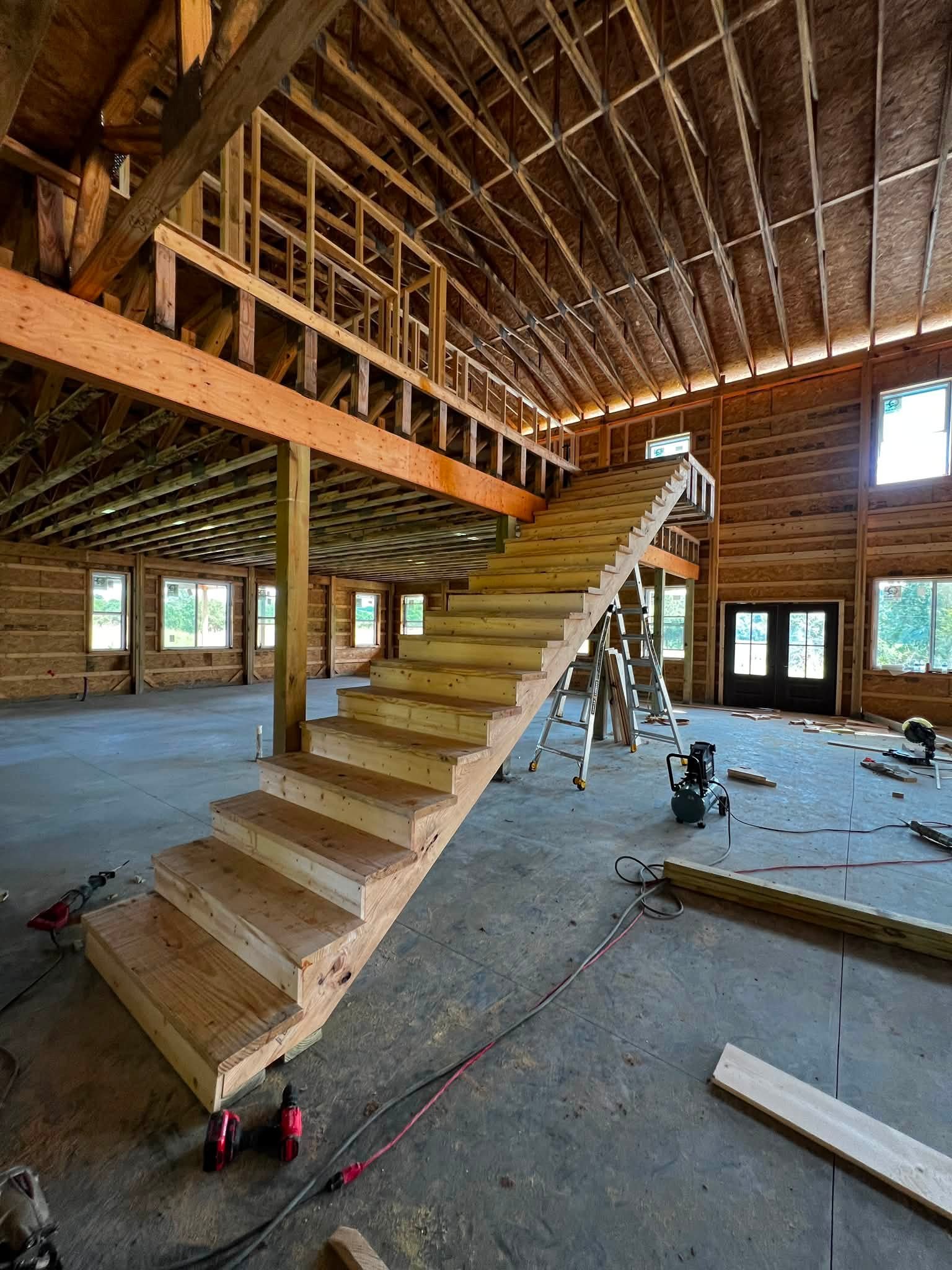 Interior view of a house under construction with exposed wooden framing, stairs, and construction tools on the floor.