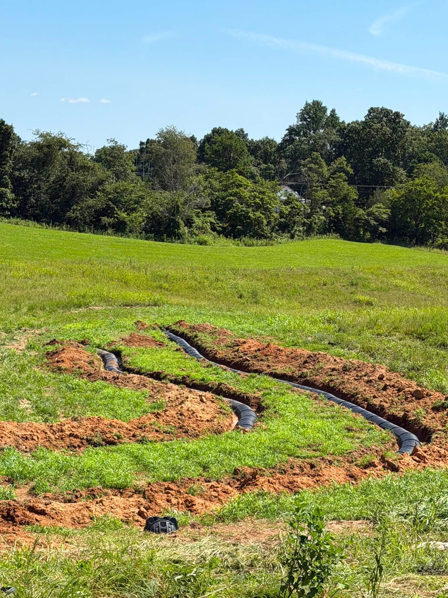 Freshly dug trenches for underground piping in a green field with trees and a house in the background, under a blue sky.