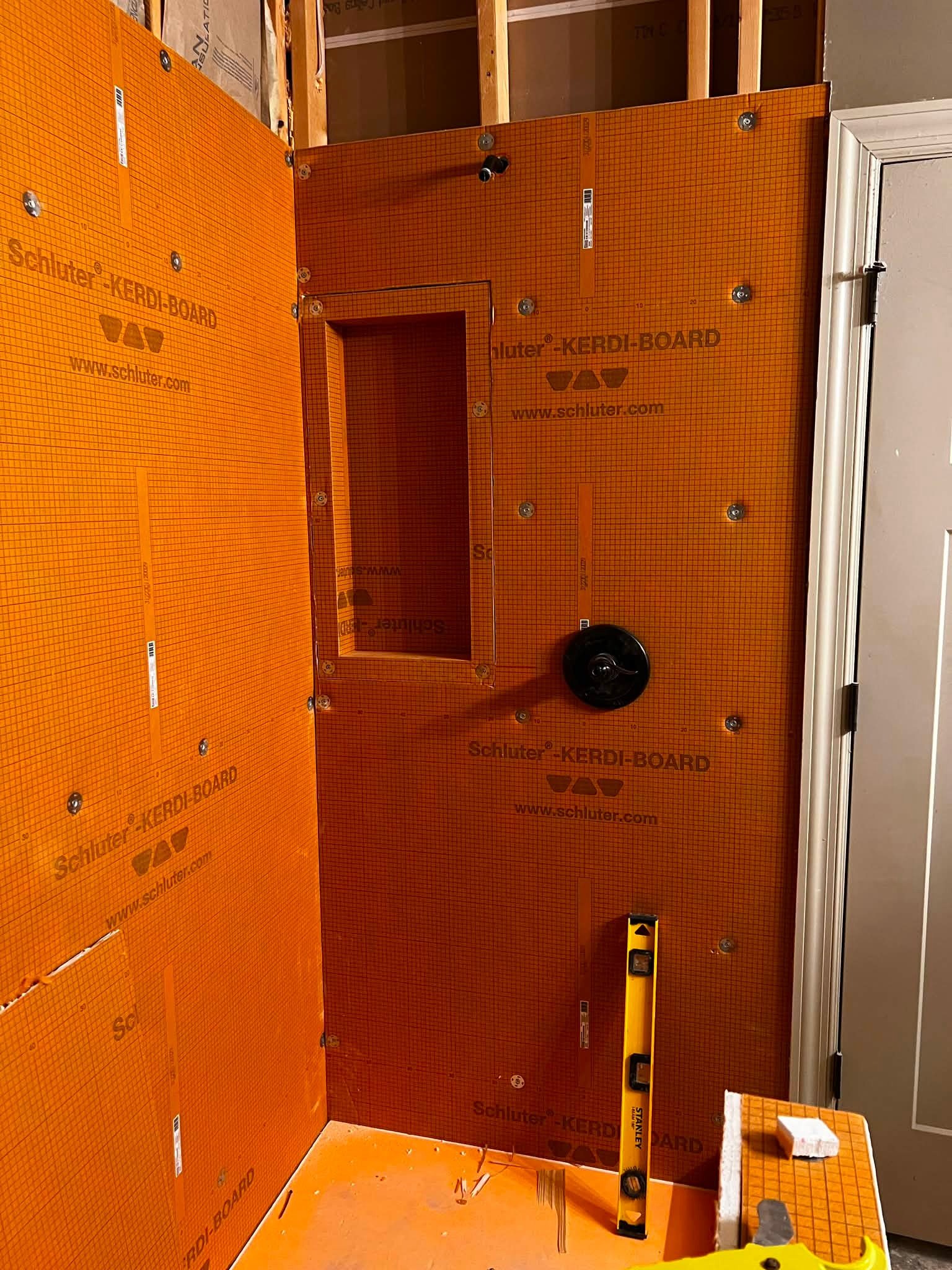 Interior view of a room under construction or renovation with orange drywall panels labeled Schlüter KERDI-BOARD, a level tool, a piece of foam, and some scattered construction debris.