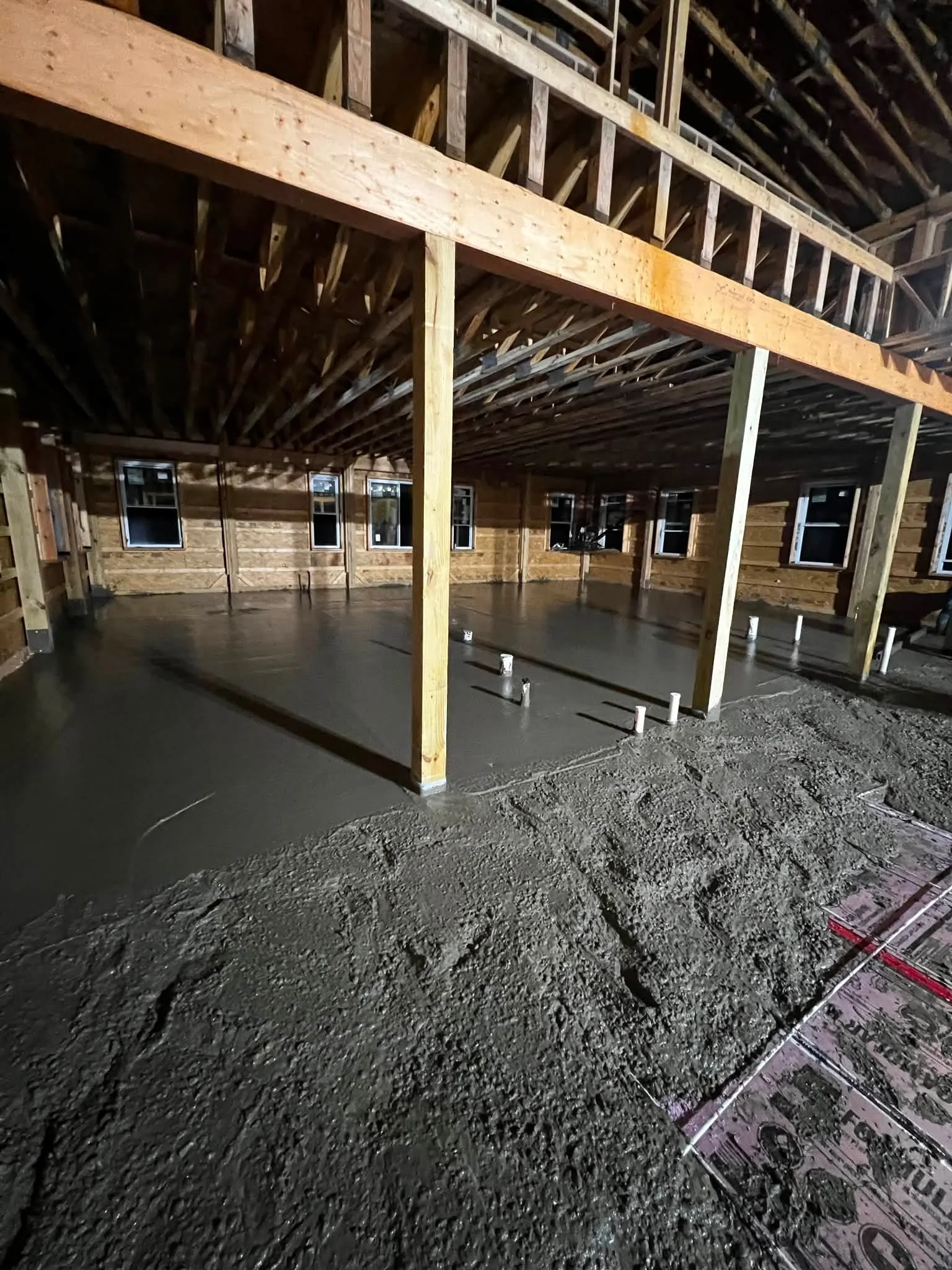 The image shows the interior of a building under construction with exposed wooden framing and a concrete floor. The space has multiple windows along the wooden wall, and construction materials and pipes are visible on the floor.