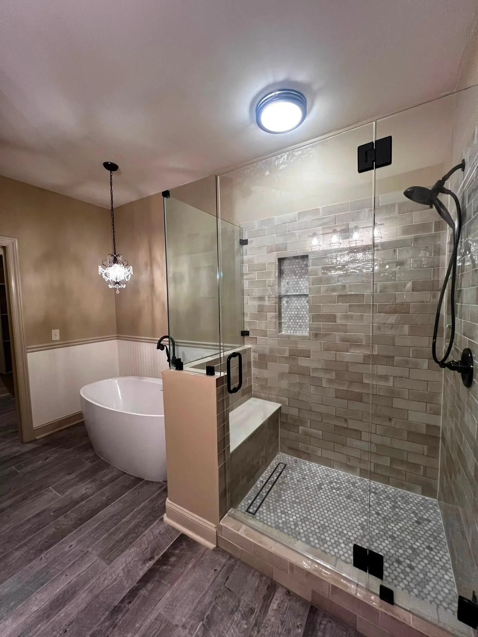 A modern bathroom with a walk-in shower enclosed by glass. The shower has beige brick-style tile walls, a built-in niche, and a pebble tile floor with a linear drain. Outside the shower, there is a white freestanding bathtub, dark wood flooring, beig