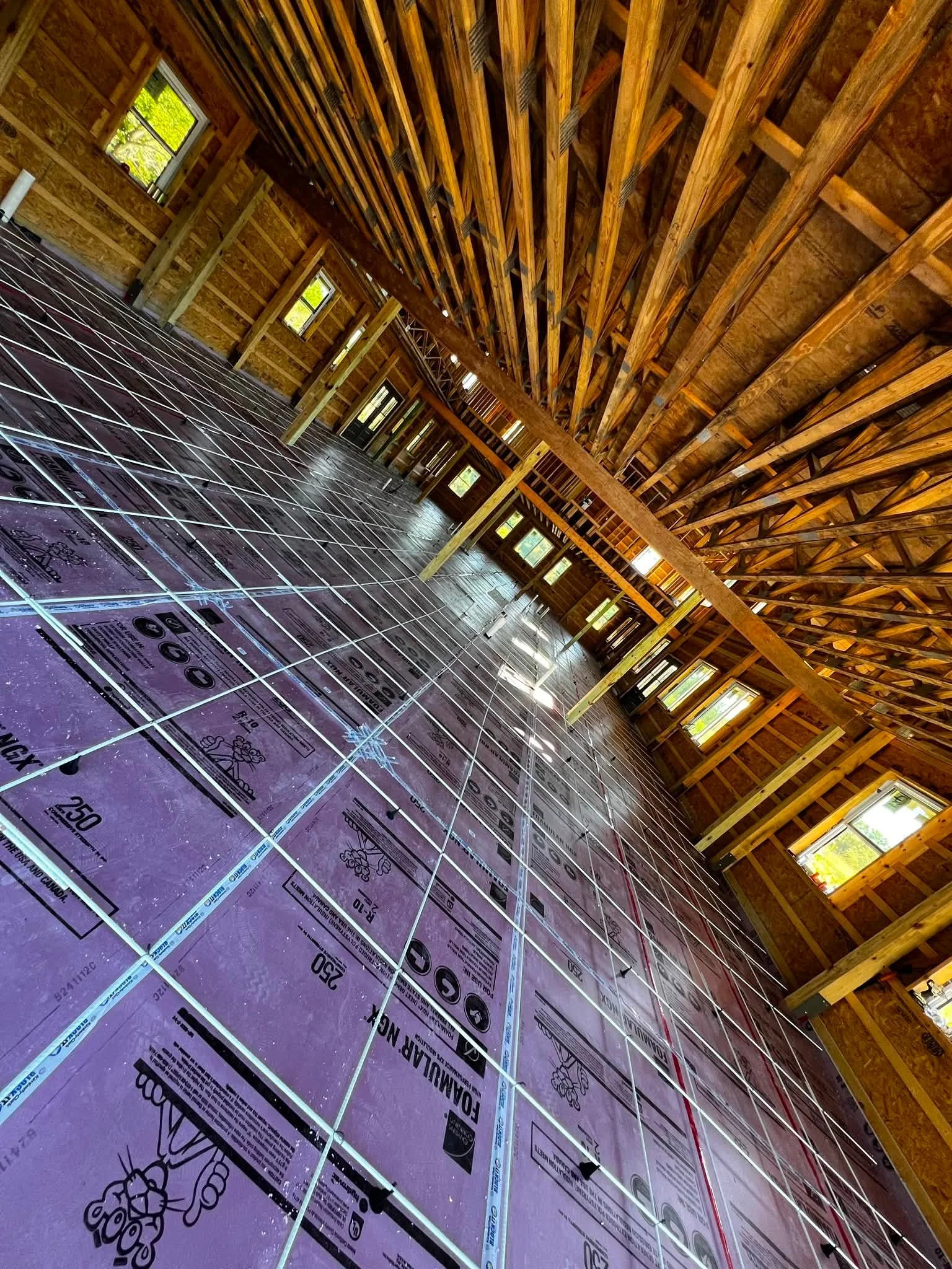 Interior of a house under construction with wooden framing and purple insulation foam sheets on the floor, multiple windows along the walls, and electrical wiring installed.