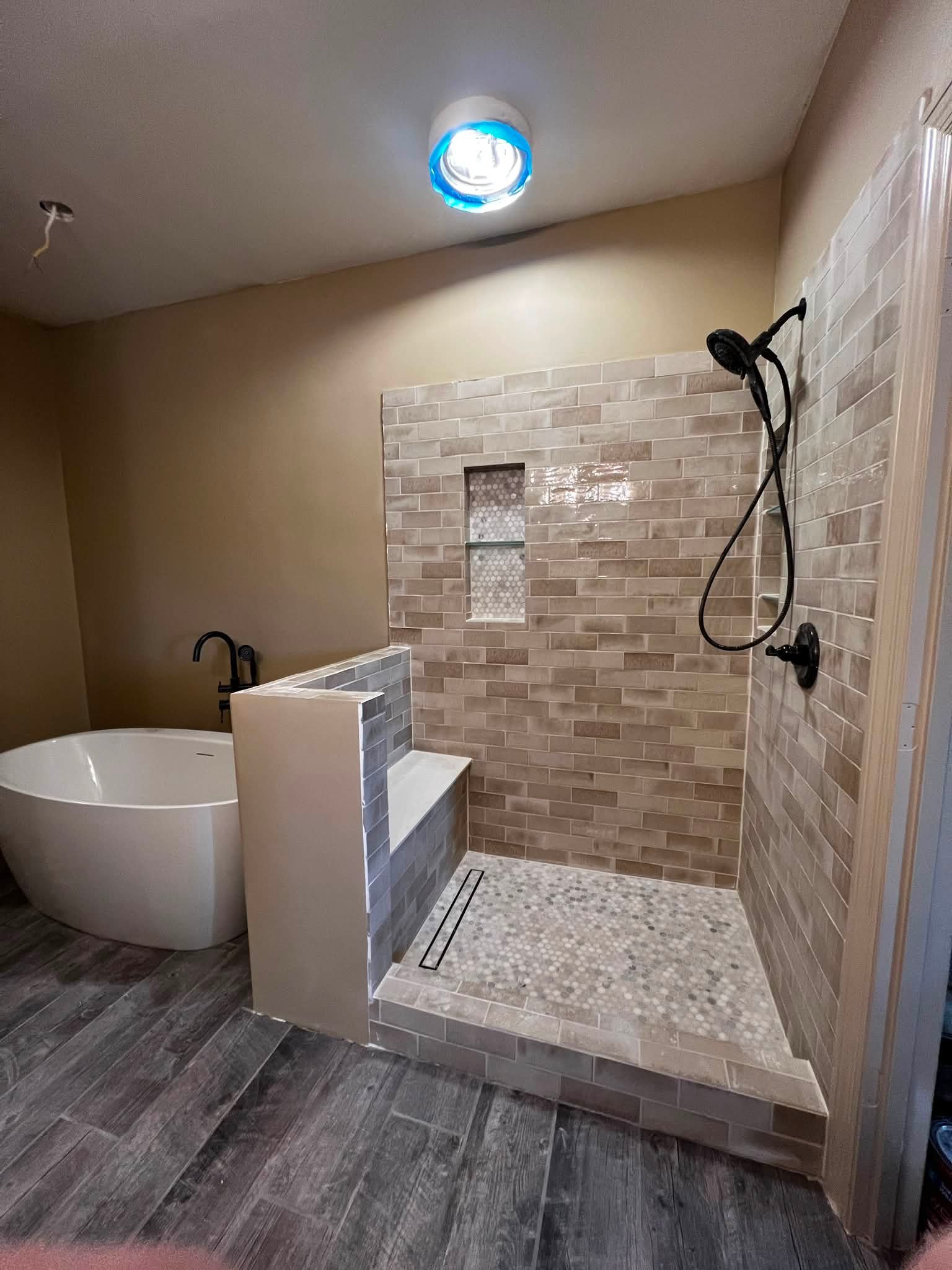A bathroom with a walk-in shower area enclosed by a half wall and tiled walls. The shower has a black handheld showerhead, a narrow built-in shelf, and a floor drain. The bathroom features wood-look floor tiles and neutral-colored walls.