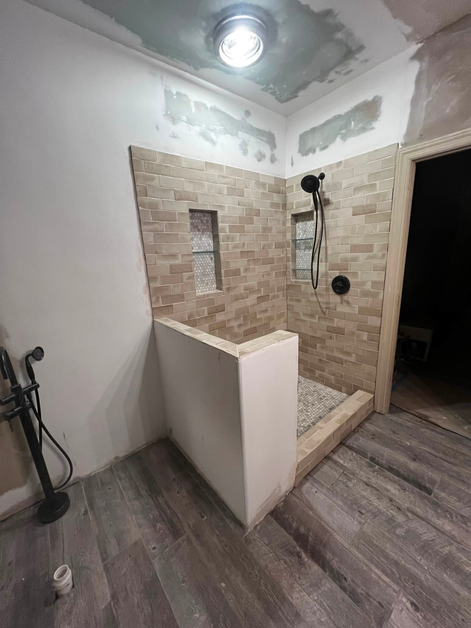 A bathroom with a shower area that has brick tile walls, a built-in niche, and a raised shower platform with mosaic tile flooring. Exposed drywall on the ceiling and part of the walls, with some unpainted and unfinished surfaces. Shower fixtures incl