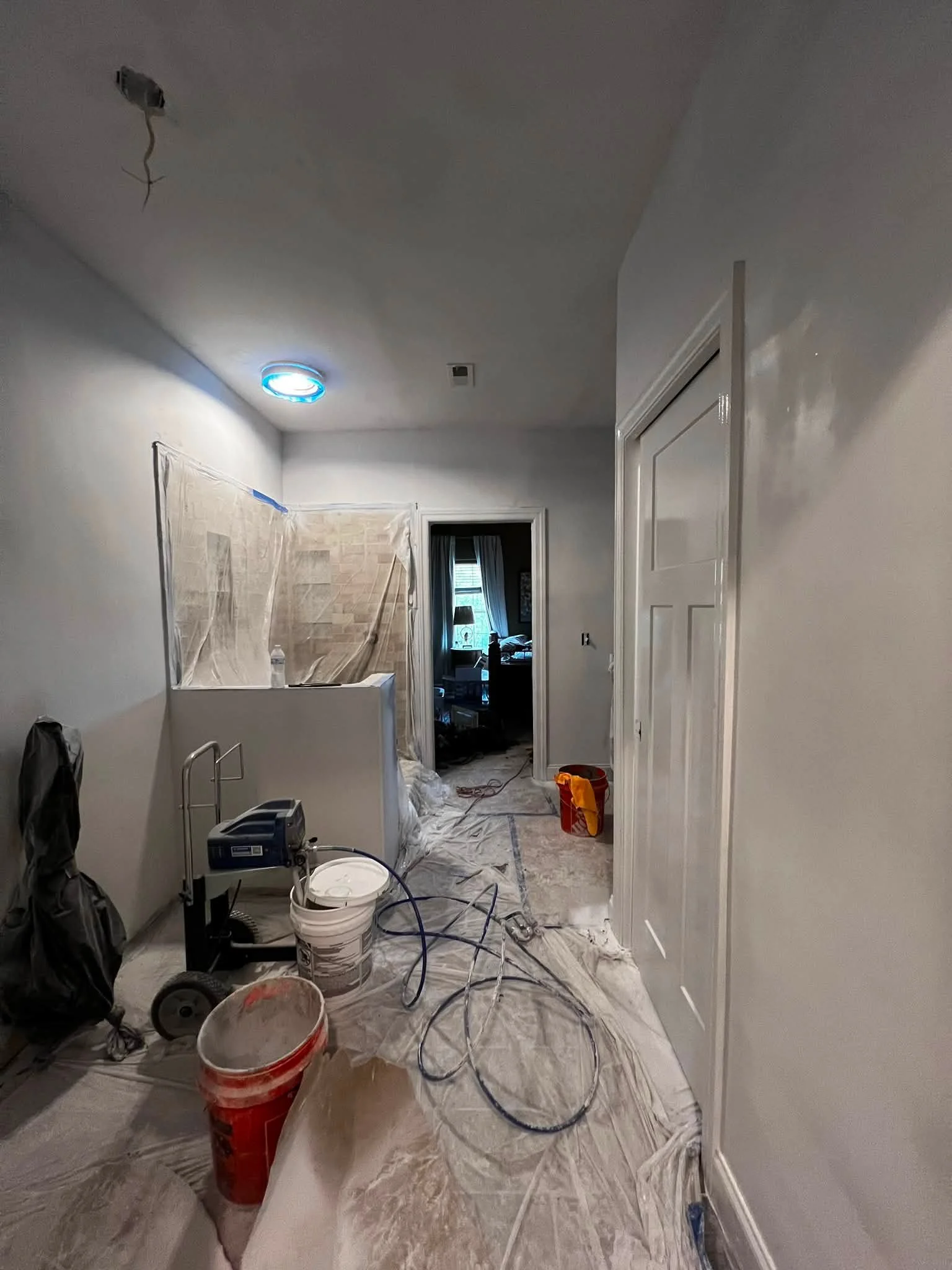 Interior room under renovation with plastic-covered furniture and painting supplies, such as paint buckets, rollers, and drop cloths on the floor. There is a doorway to another room with a window and curtains visible in the background.