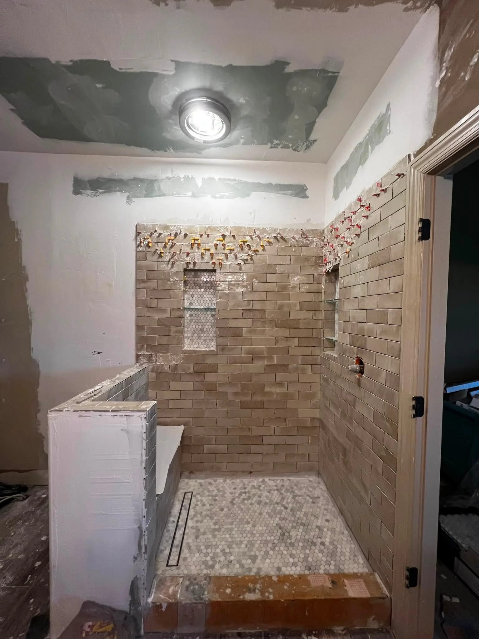 A bathroom under renovation, showing partially tiled walls with brick and stone tiles, a built-in niche with small mosaic tiles, a partially installed shower pan with hexagonal tiles, and construction tools and materials around.